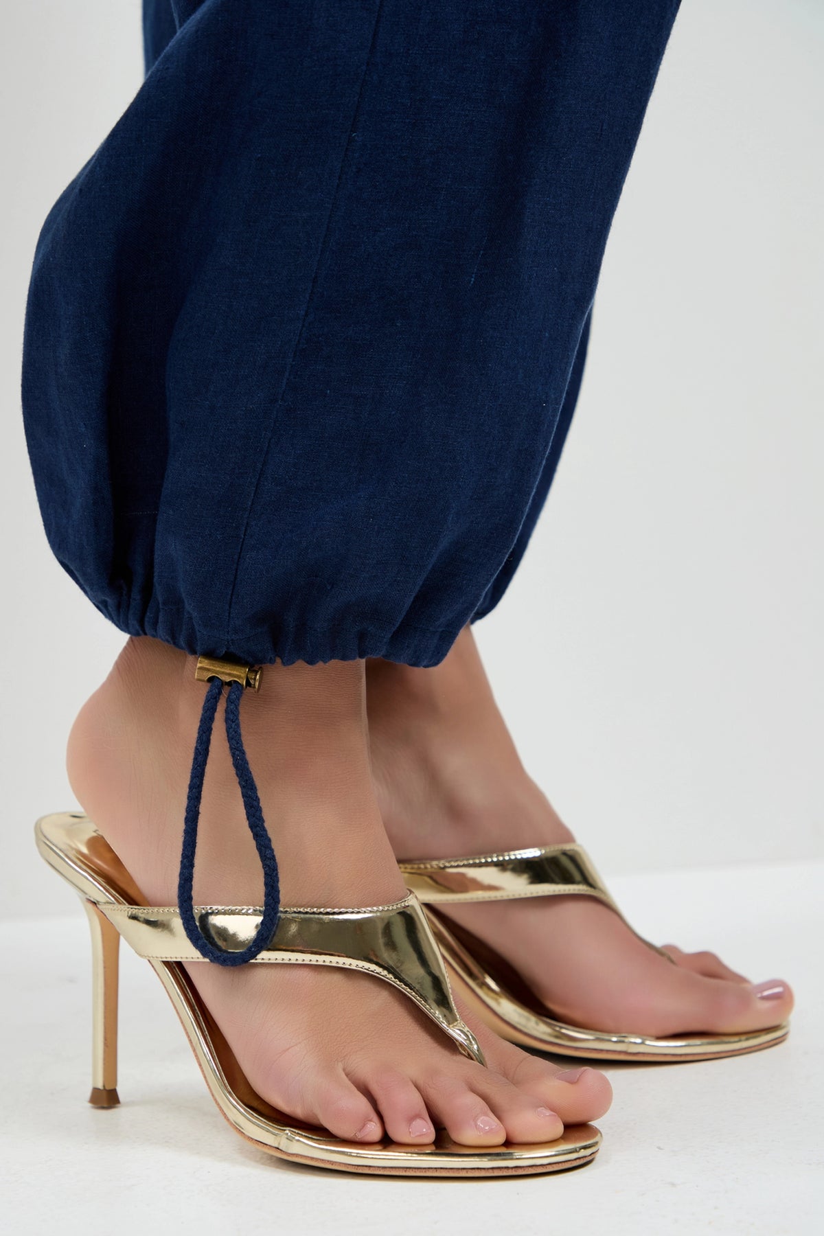 A person wearing gold high-heeled sandals and navy blue Alico Cargo Pants with an elastic drawstring hem, standing on a white surface.