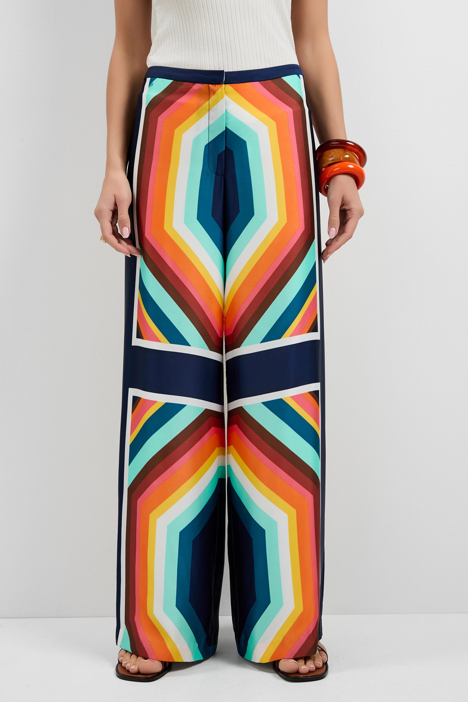 A person wears the Lanay Siena Wide-Leg Pants, featuring a bold, colorful geometric pattern in orange, blue, yellow, and red, paired with a white top and brown sandals.