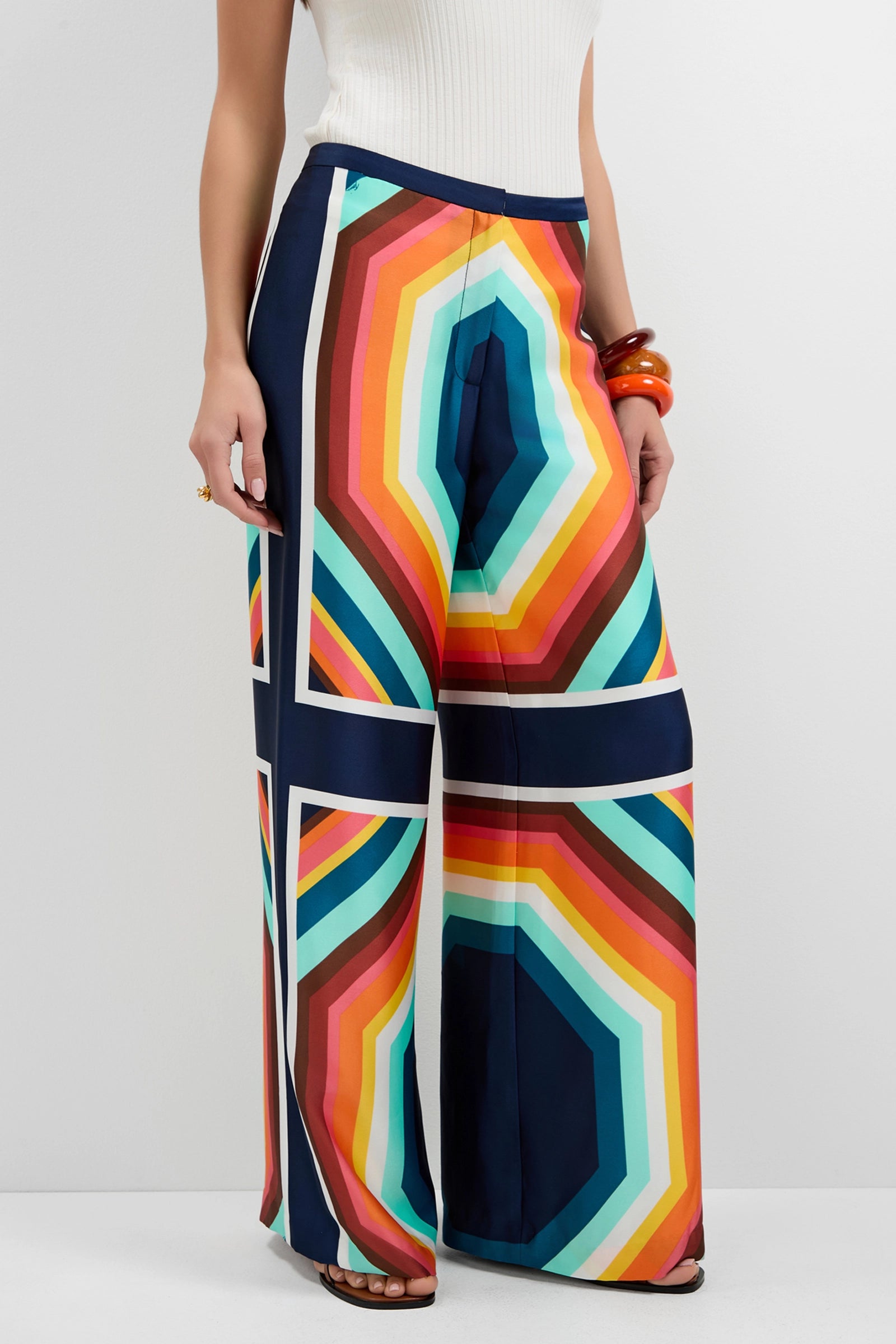 A person wears Lanay Siena Wide-Leg Pants, featuring a bold geometric pattern with hexagonal shapes in red, yellow, blue, and orange, paired with a sleeveless white top.