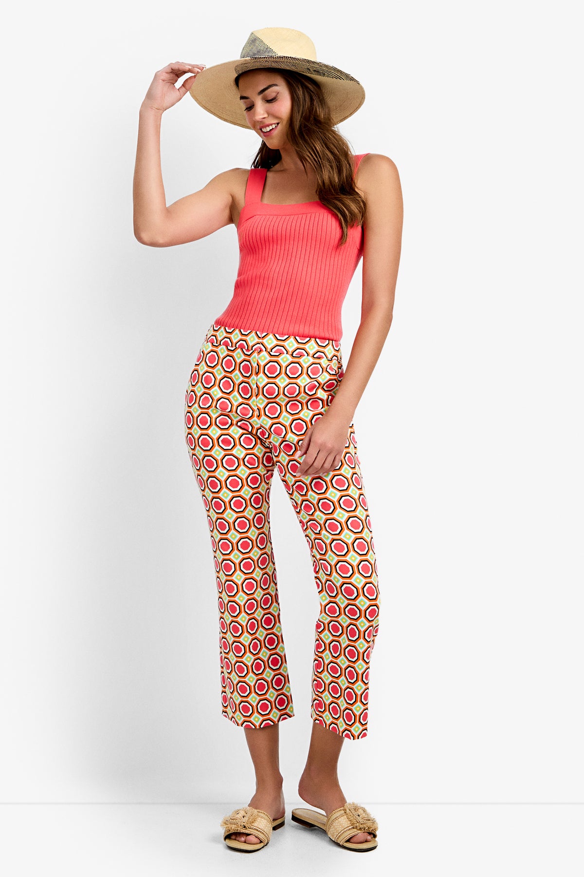 A woman smiles and poses against a white background, wearing a wide-brim straw hat, coral ribbed tank top, beige slide sandals, and Lulu Cropped Pants - Salerno with red and yellow circle patterns.