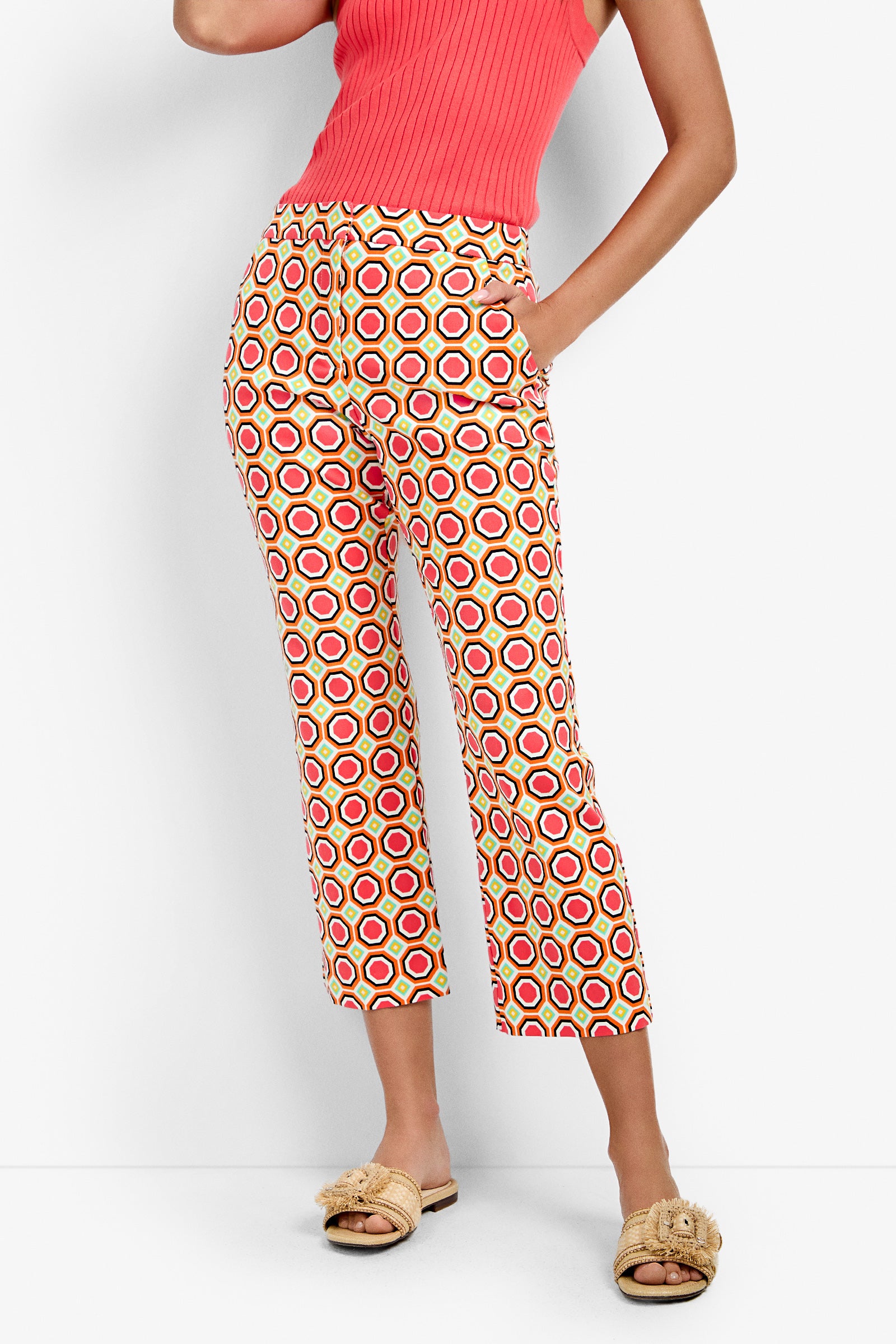 Wearing Lulu Cropped Pants - Salerno with a red and yellow circular motif, a sleeveless ribbed coral top, and beige slip-on sandals, a person stands against a white background with one hand in their pocket.