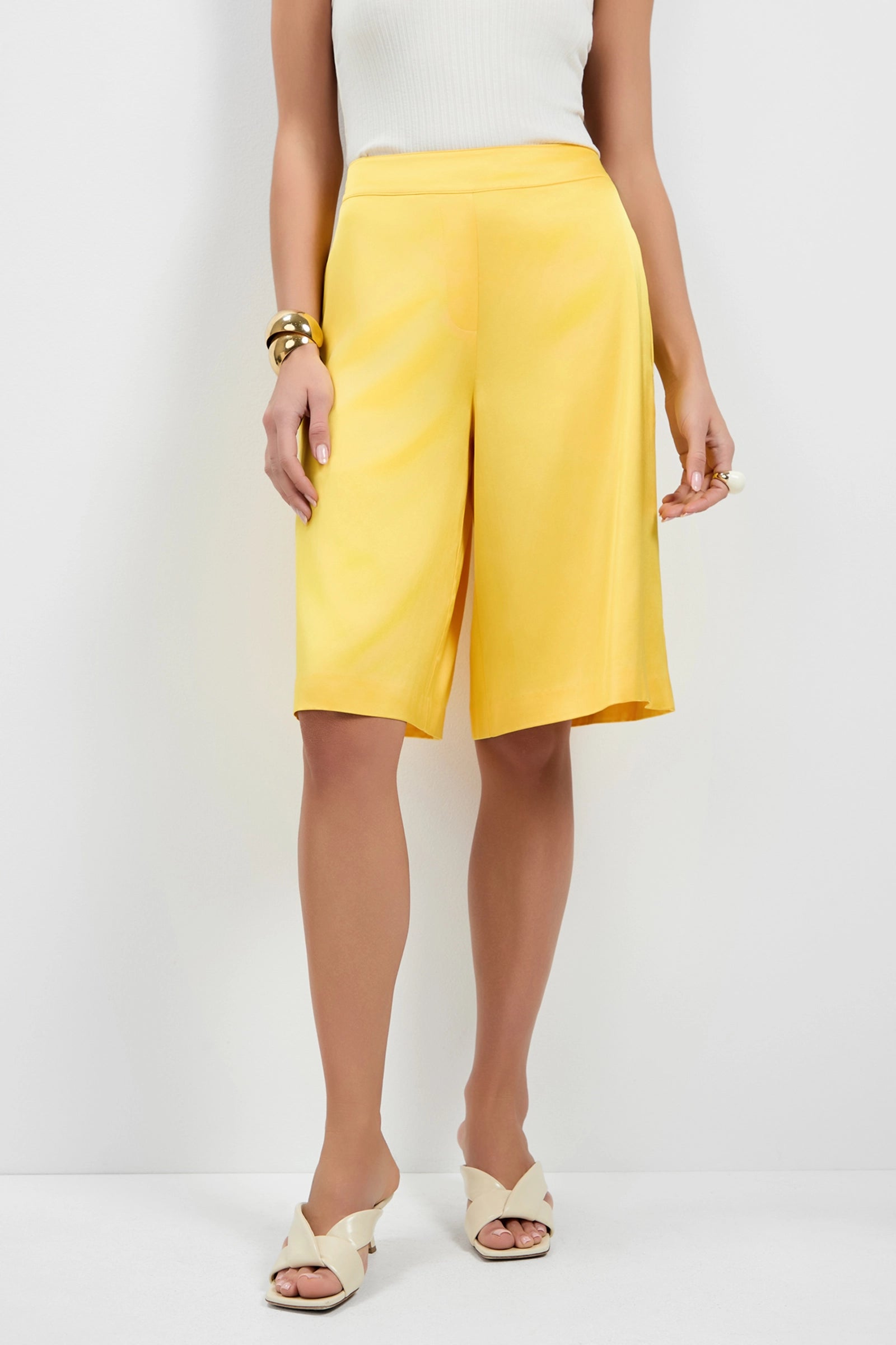 A woman wears Chiana Satin Shorts in yellow with a white sleeveless top, cream open-toe heels, and gold jewelry, standing against a plain white background. Her face is not visible.