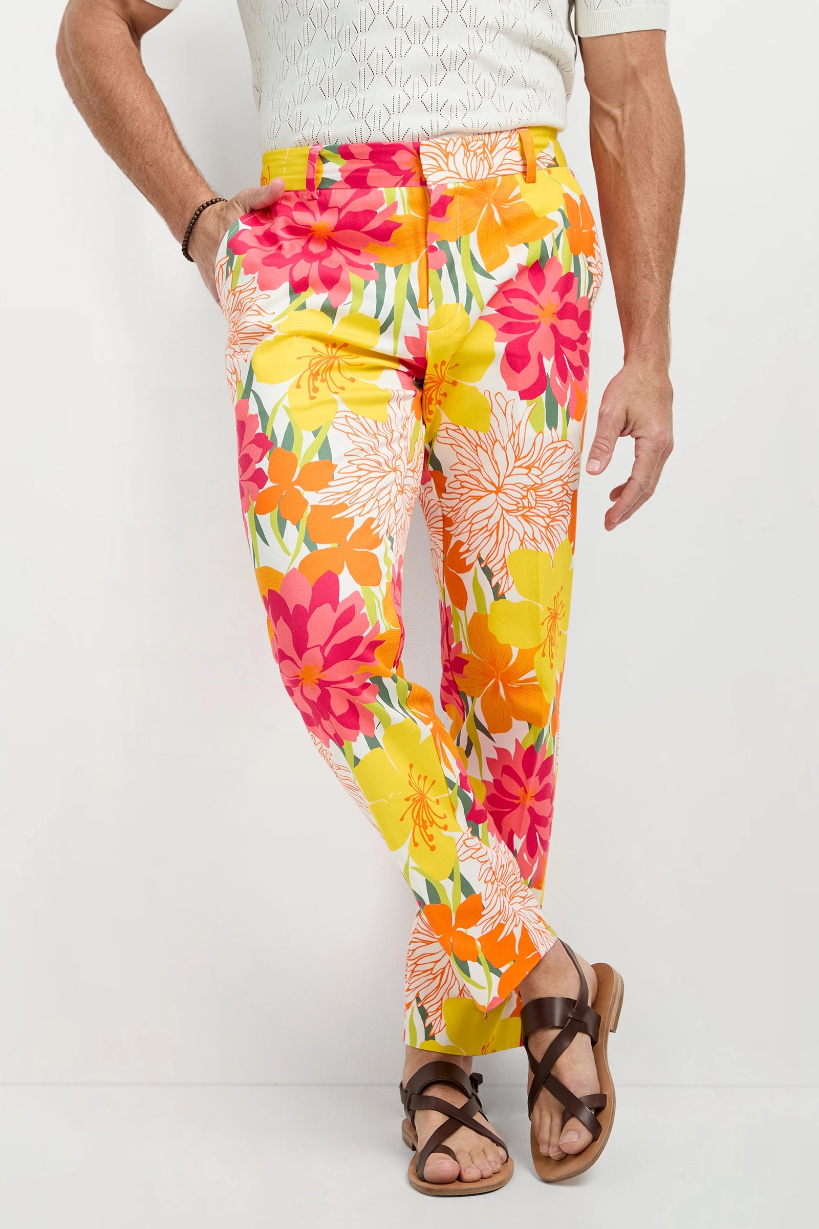 A person wears the Clyde Slim Trouser - Como Herringbone, featuring a colorful floral pattern in yellow, orange, pink, and white, paired with brown sandals and a short-sleeve shirt while standing cross-legged.
