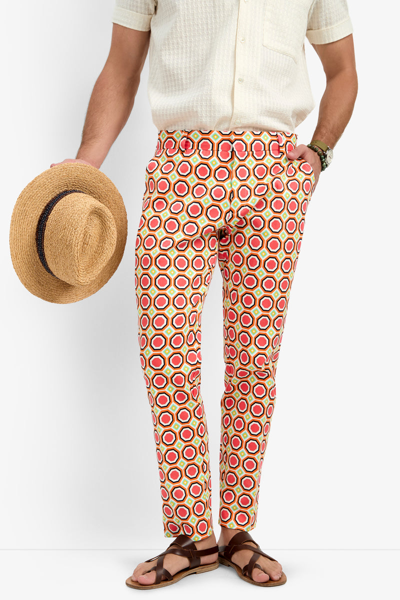 A person wearing a light short-sleeve shirt, Clyde Slim Trouser - Salerno Herringbone in a bold red, orange, and yellow circle pattern, brown sandals, and holding a straw hat by their side. The person’s head is out of frame.