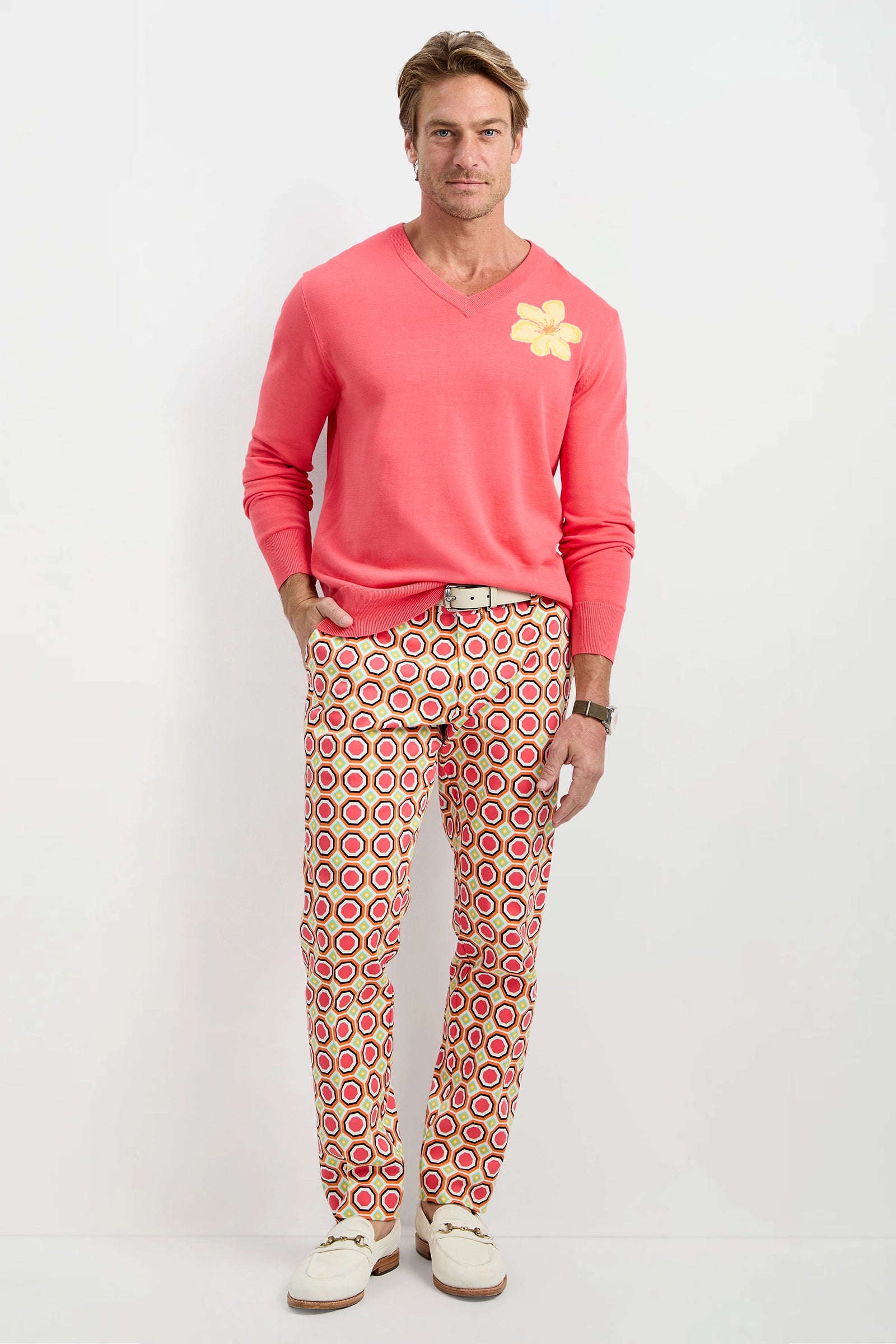 A man stands against a plain white background wearing a pink sweater with a yellow flower, Clyde Slim Trouser - Salerno Herringbone featuring red, yellow, and white circle patterns, and white shoes. One hand is in his pocket.