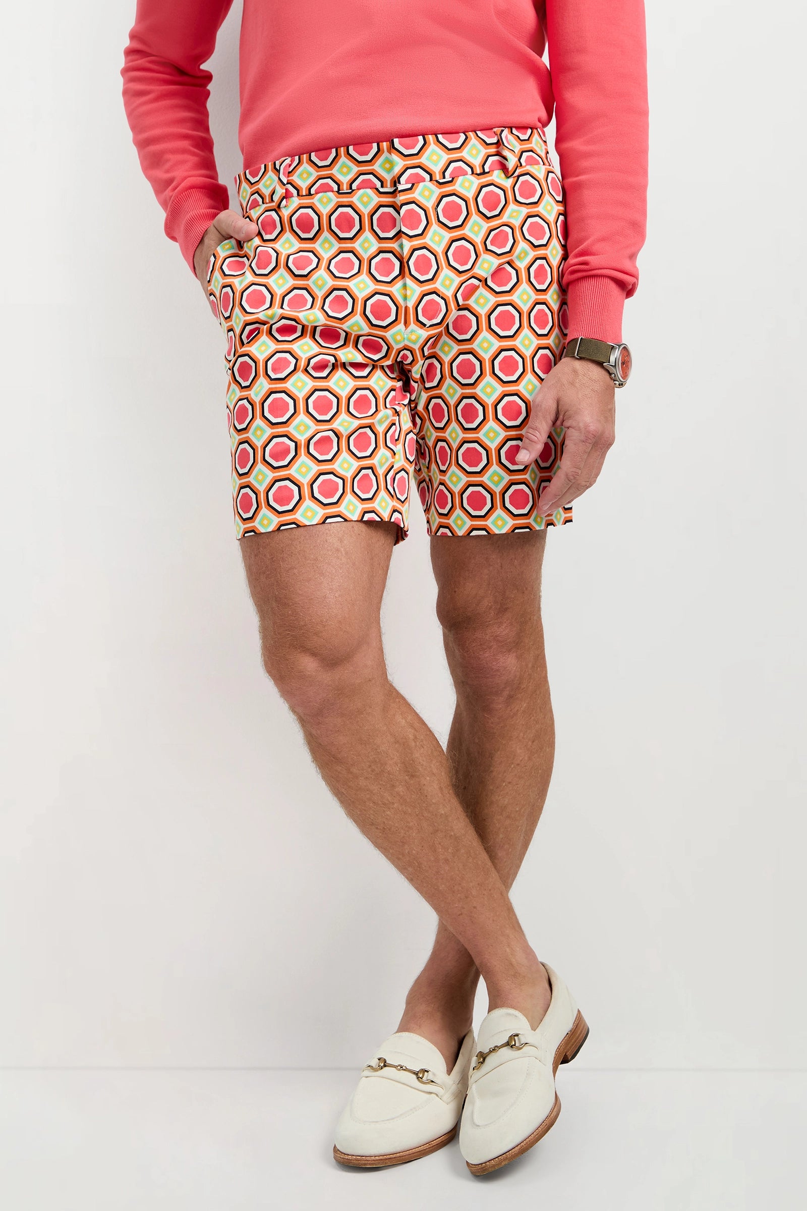 A man stands with hands in pockets on a light background, wearing a coral sweatshirt, the Lawrence Short - Salerno Herringbone, a watch, and cream loafers.