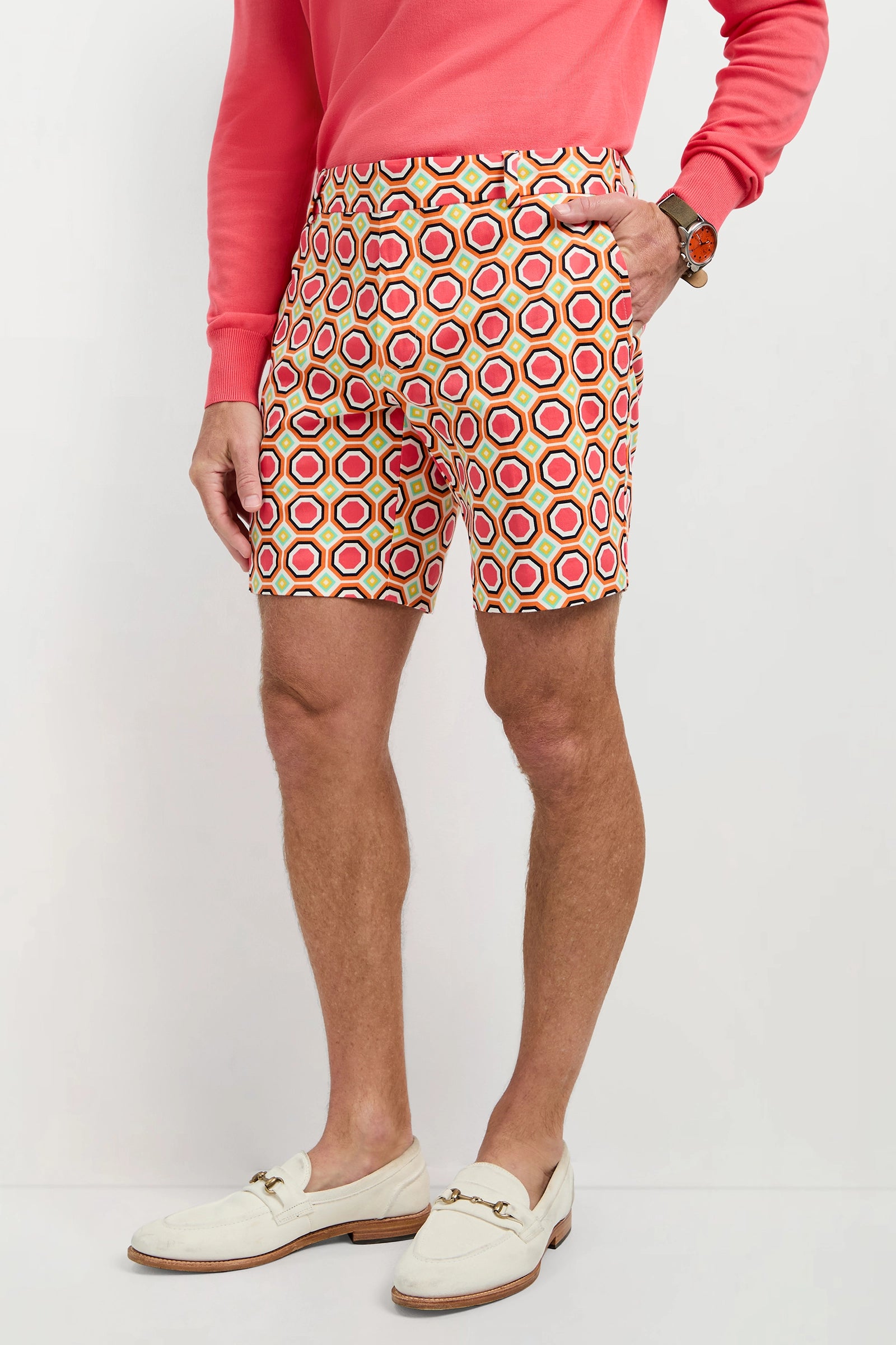 A person wearing a coral long-sleeve shirt, Lawrence Short - Salerno Herringbone with a hexagon pattern, a brown watch, and white loafers stands against a plain background.