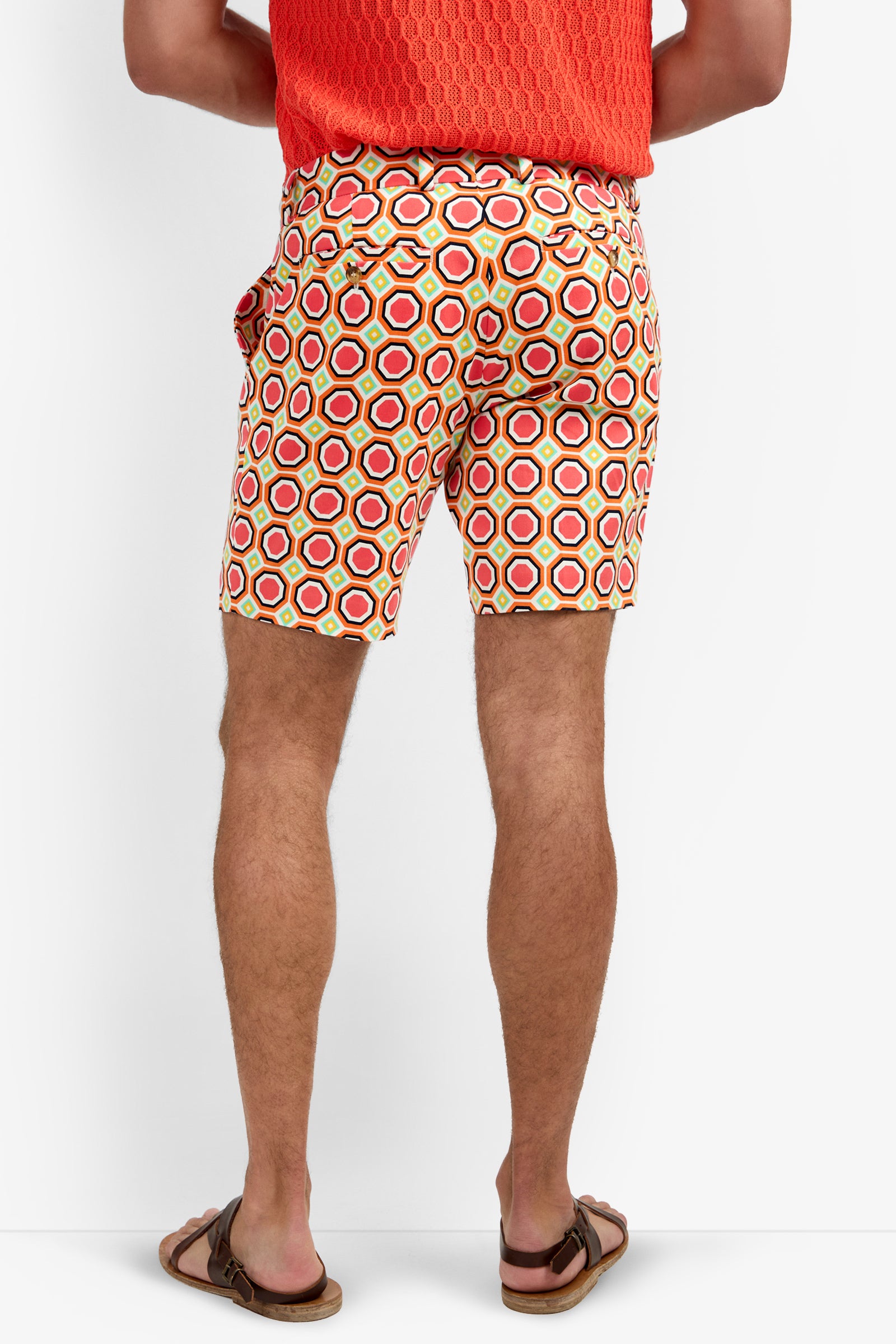 Wearing the Lawrence Short - Salerno Herringbone, a person is shown from the lower back against a white background, paired with a bright orange textured shirt and brown sandals. The shorts feature a red, yellow, and white geometric pattern.