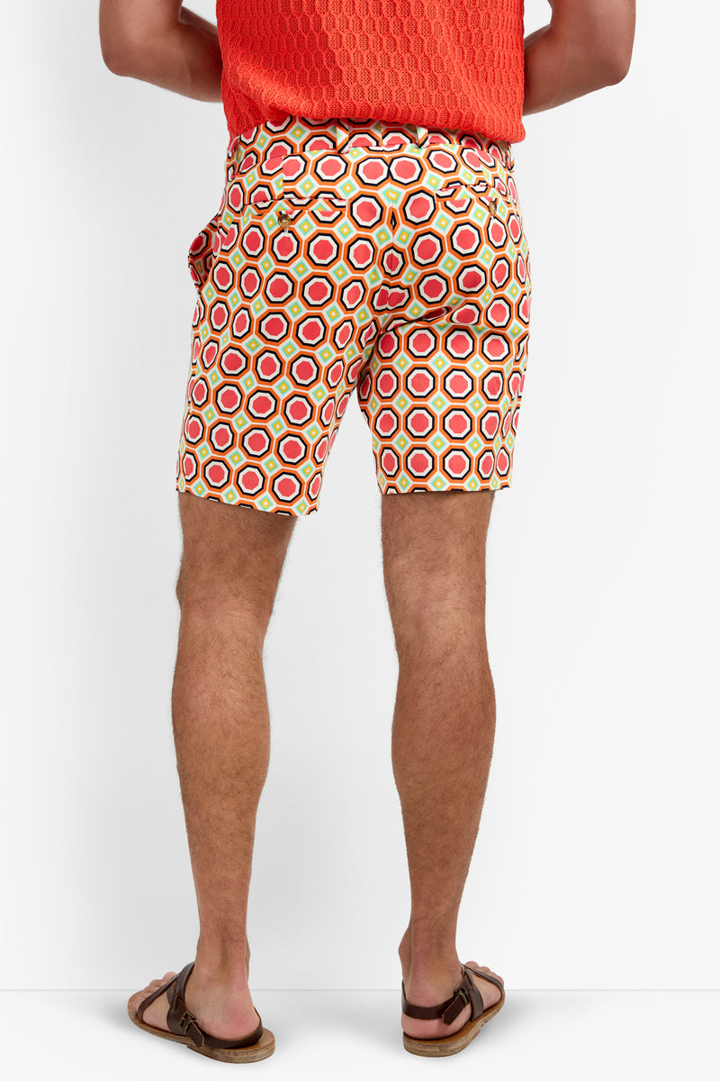 Wearing the Lawrence Short - Salerno Herringbone, a person is shown from the lower back against a white background, paired with a bright orange textured shirt and brown sandals. The shorts feature a red, yellow, and white geometric pattern.