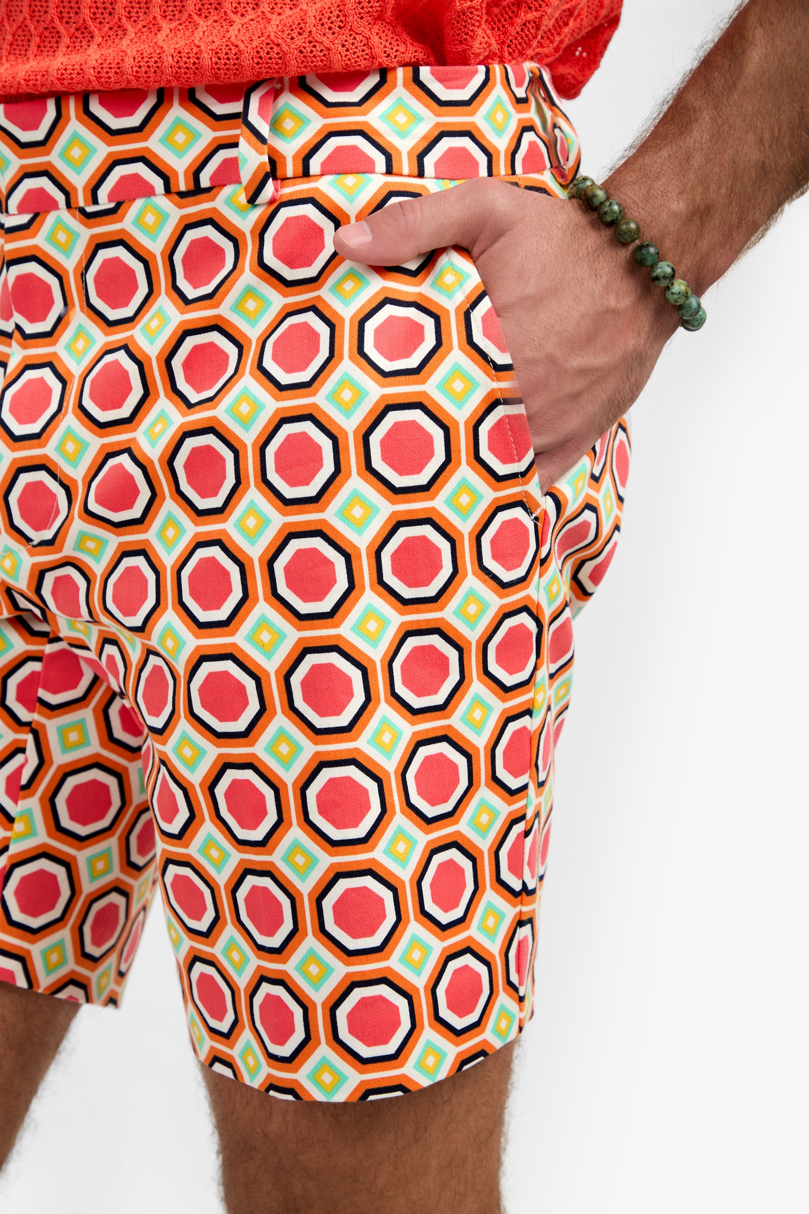 A person wearing the Lawrence Short - Salerno Herringbone, featuring colorful geometric patterns, paired with a coral shirt and a green beaded bracelet, stands with one hand in their pocket.
