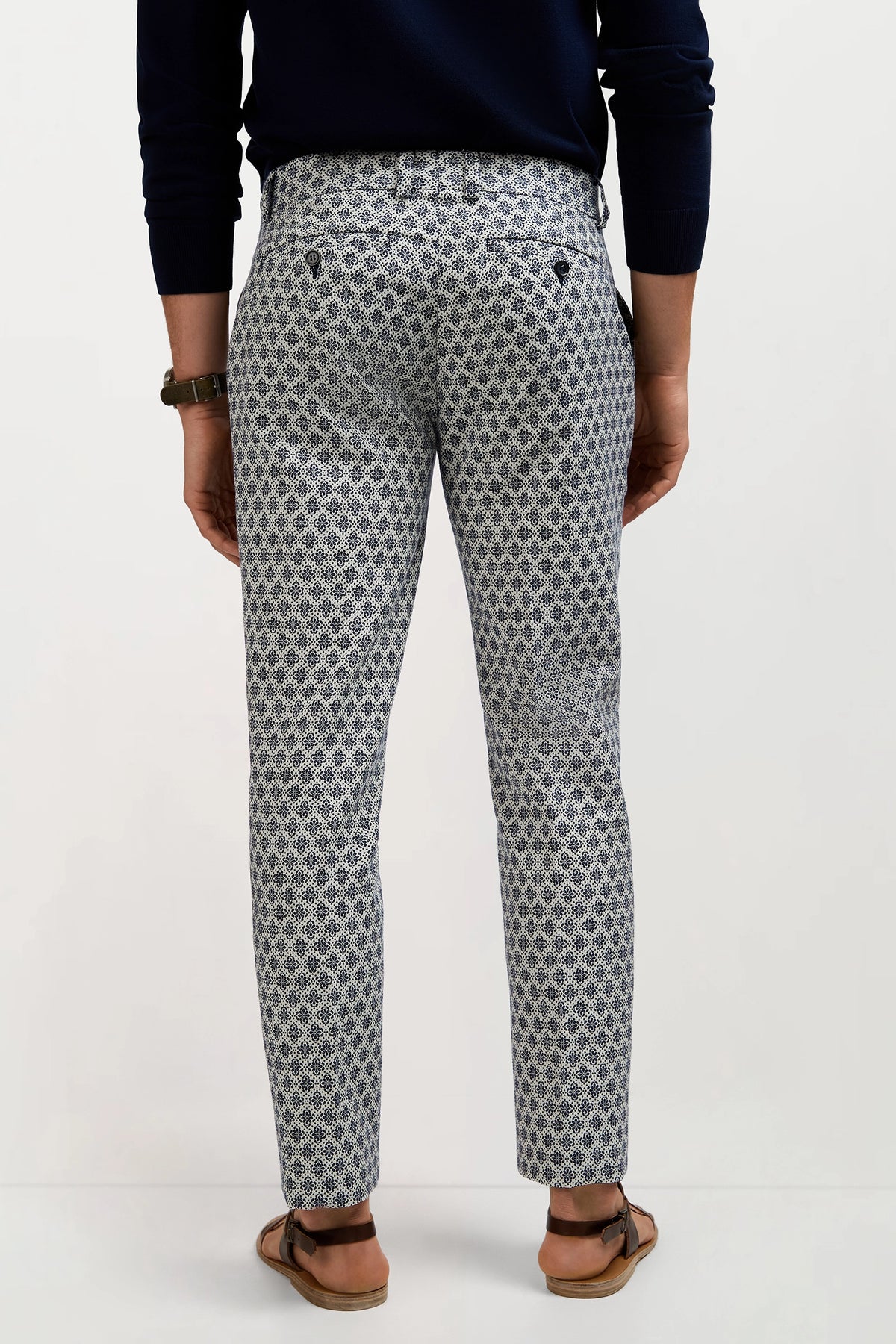 A person stands facing away, wearing the Clyde Slim Trouser - Daisy Jacquard with a dark long-sleeve top, sandals, and a watch against a plain light background.