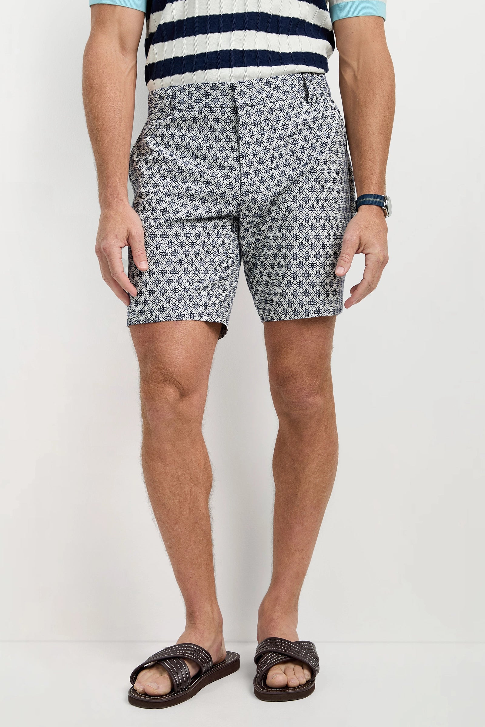 A man wearing the Lawrence Short - Daisy Jacquard stands against a plain light background. He also wears a striped short-sleeve sweater, wristwatch, and brown woven sandals. His head is not visible in the image.