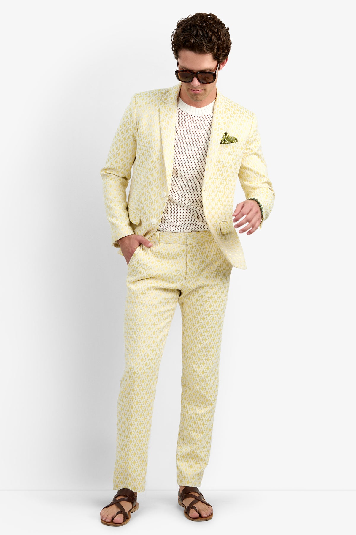 A man wears the Clyde Slim Trouser - Crossroads Jacquard with a light yellow patterned suit, white mesh shirt, brown sandals, green pocket square, and sunglasses, standing against a plain background and looking down.