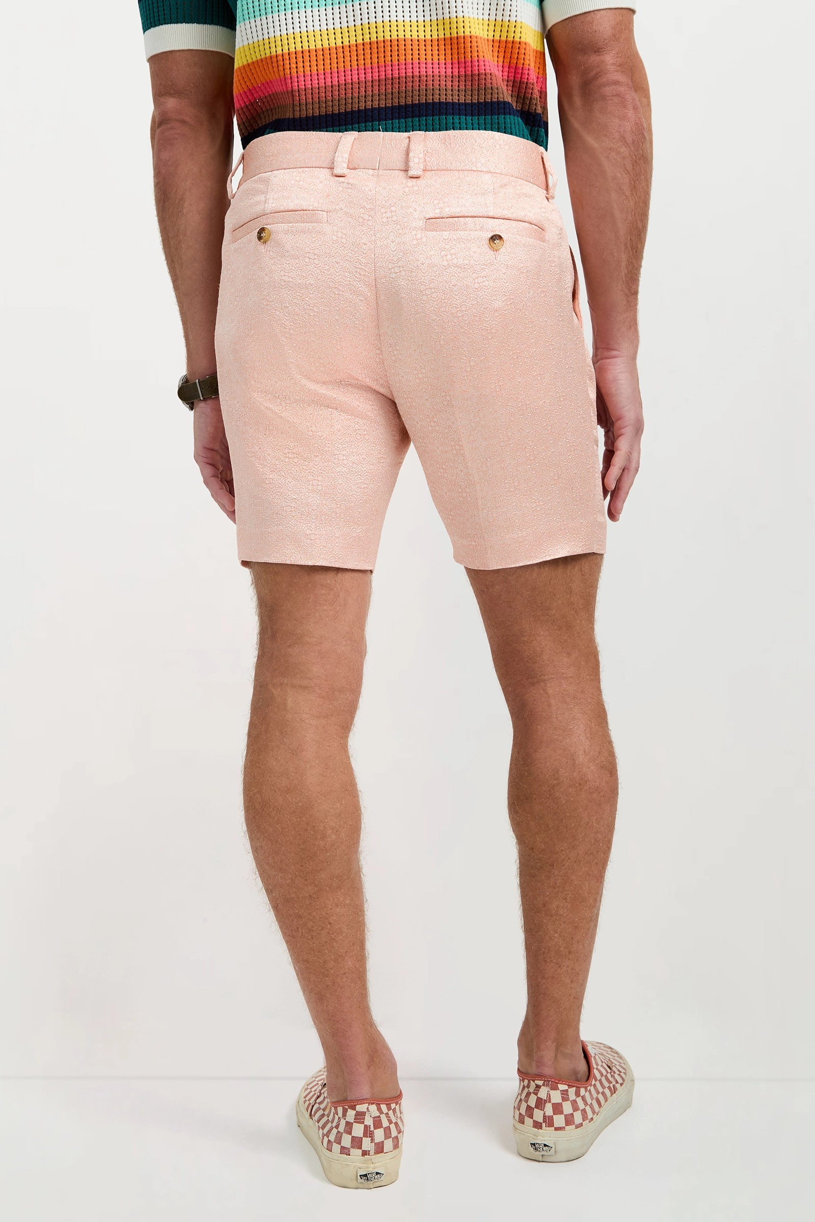 A person stands facing away, wearing the Clyde Short - Metallic Jacquard in light pink, paired with a colorful striped shirt and red and white checkered slip-on shoes. Their arms are relaxed at their sides.