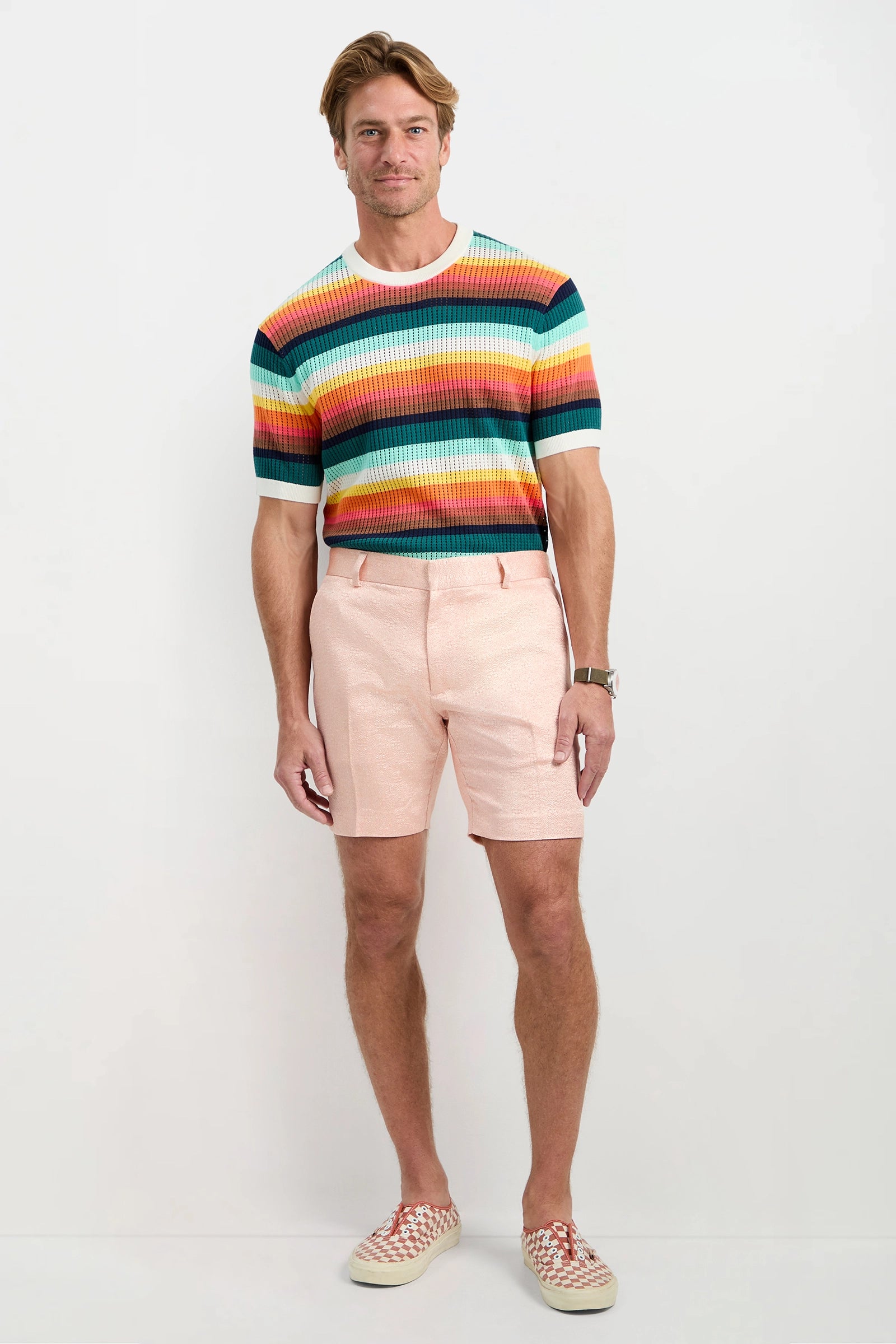 A man stands against a plain white background smiling and wearing the Clyde Short - Metallic Jacquard, paired with a colorful, striped short-sleeve sweater and checkerboard slip-on shoes. One hand is by his side, the other relaxed.