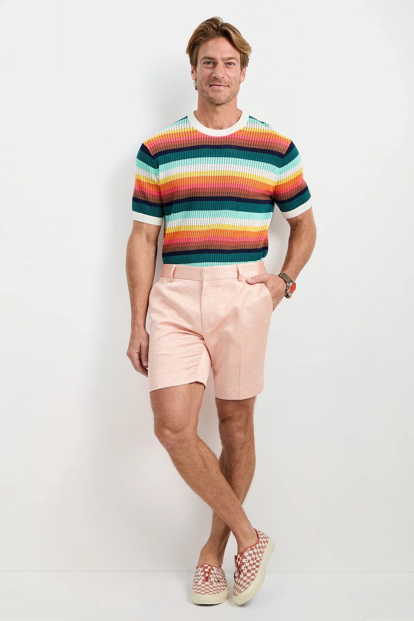 A man poses against a white wall wearing the Clyde Short - Metallic Jacquard, pairing it with a colorful striped sweater and slip-on shoes. He smiles with one hand in his pocket and the other relaxed by his side.