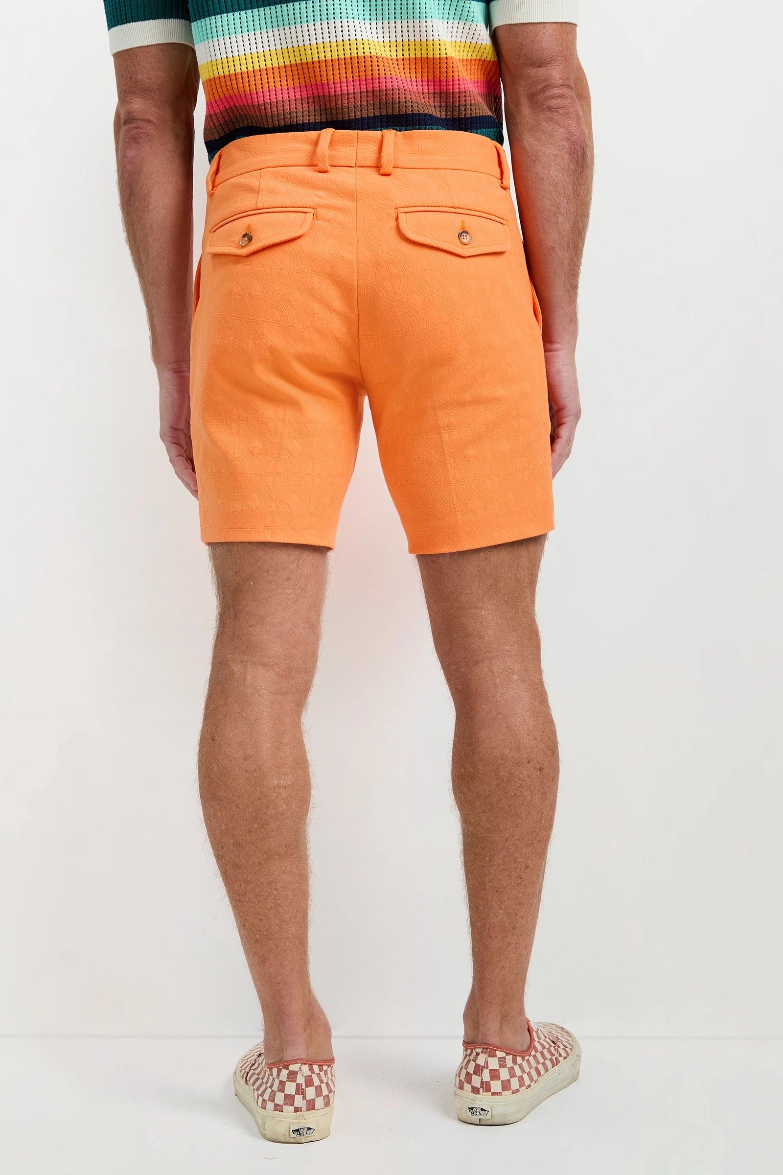 A man stands facing away, wearing the Travis Short - Tegola Jacquard with buttoned back pockets, a colorful striped shirt, and checkered slip-on shoes against a plain background.