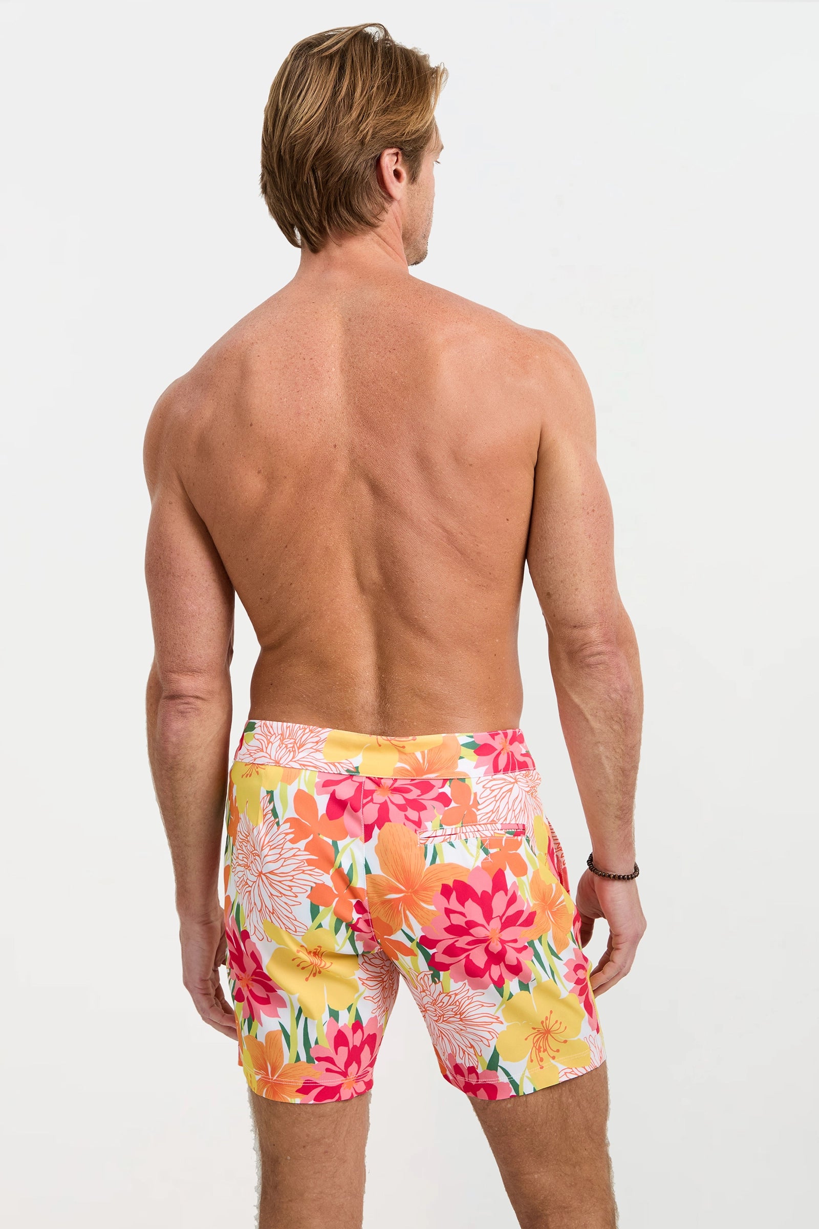 A man with light brown hair stands shirtless, facing away, wearing the Honolulu Board Short - Como in vibrant yellow, orange, pink, and red floral print against a plain white background.