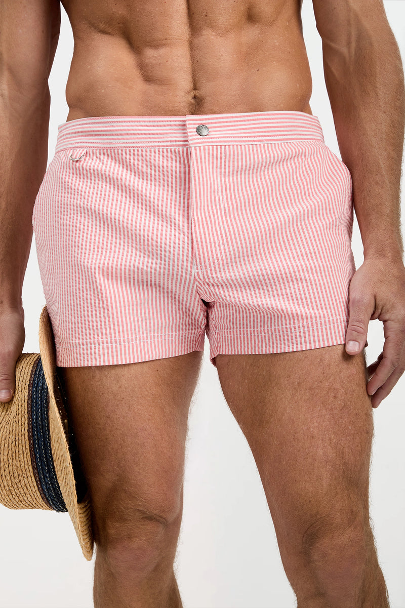 A man models the Chico Swim Trunk - Seersucker in pink and white stripes, holding a straw hat by his side. The image highlights his torso and upper legs against a plain background.