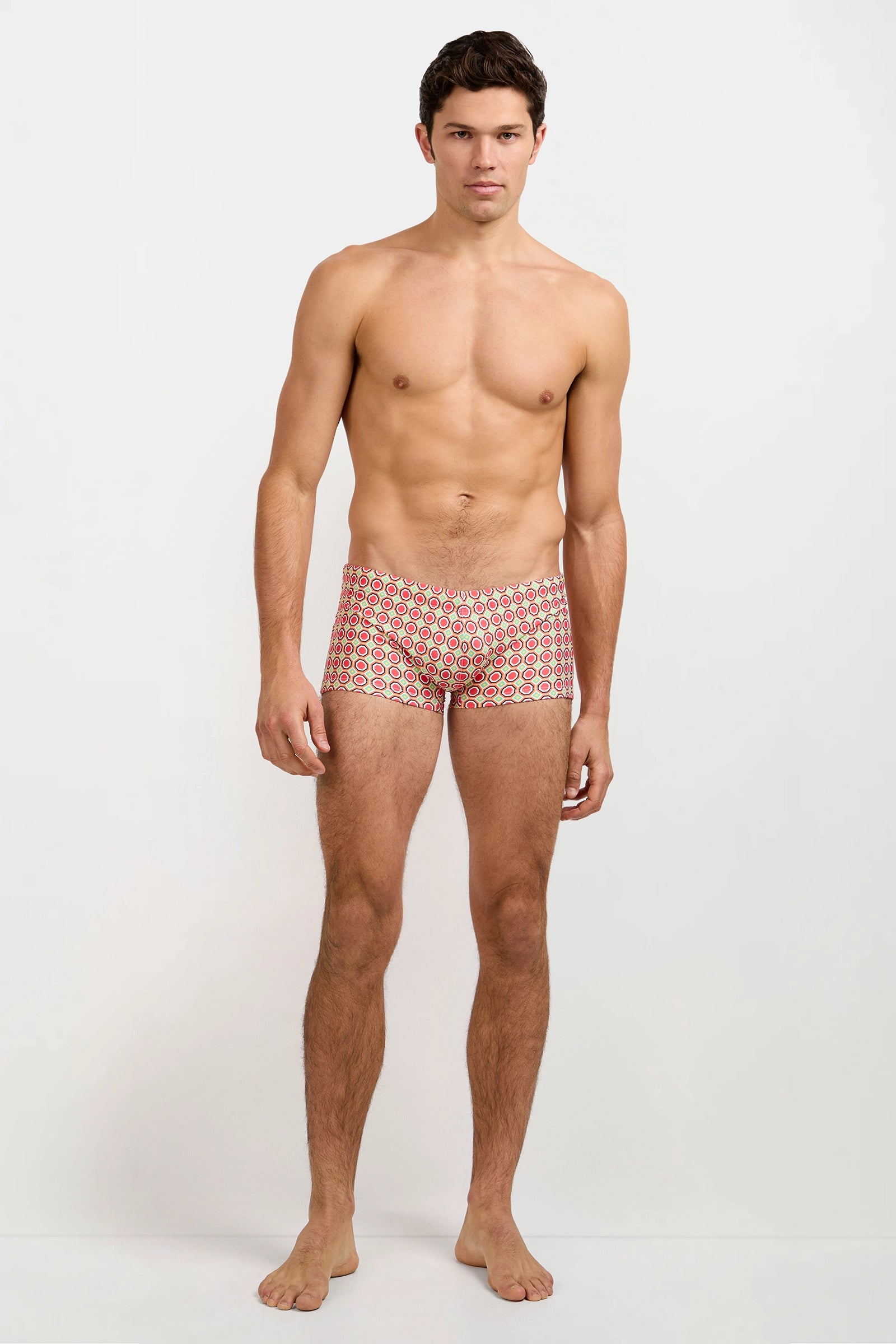 A man stands barefoot against a plain white background, wearing the Montego Bay Swim Trunk - Salerno, which features a red and white geometric pattern. He faces forward with a neutral expression and his arms relaxed at his sides.
