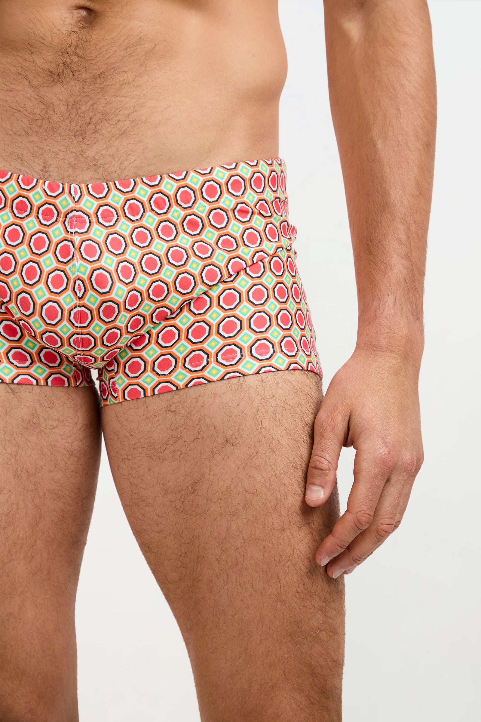 A close-up of a man's lower torso and upper thighs wearing the Montego Bay Swim Trunk - Salerno, featuring colorful geometric patterns; his right arm rests by his side against a plain white background.