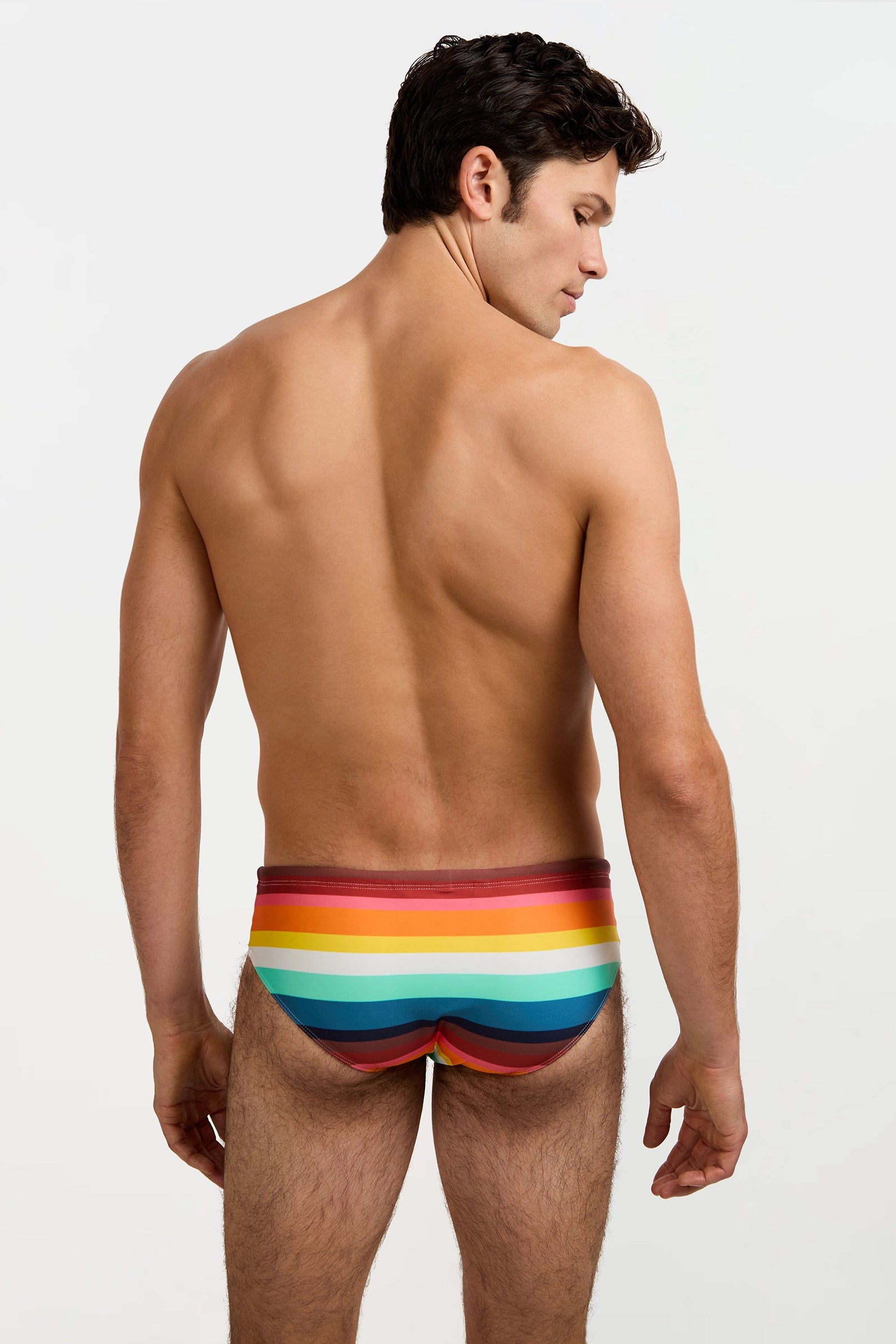 A man stands with his back to the camera, wearing the Bondi Swim Brief in Siena Stripe, featuring vibrant rainbow stripes against a plain white background.