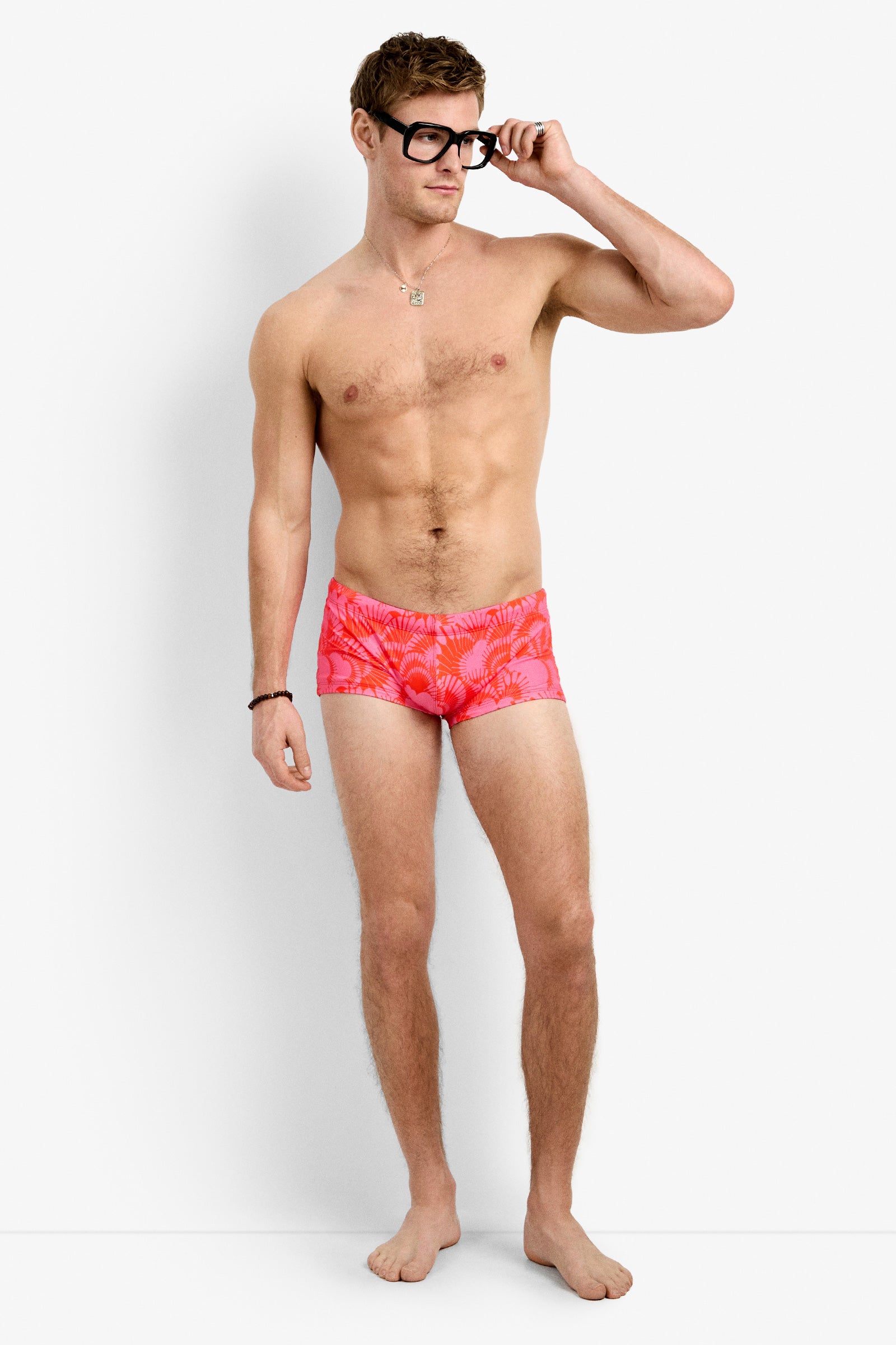 A shirtless man in large black glasses and bright Sorrento Swim Trunk stands barefoot against a plain white background, looking to the side while adjusting his glasses.