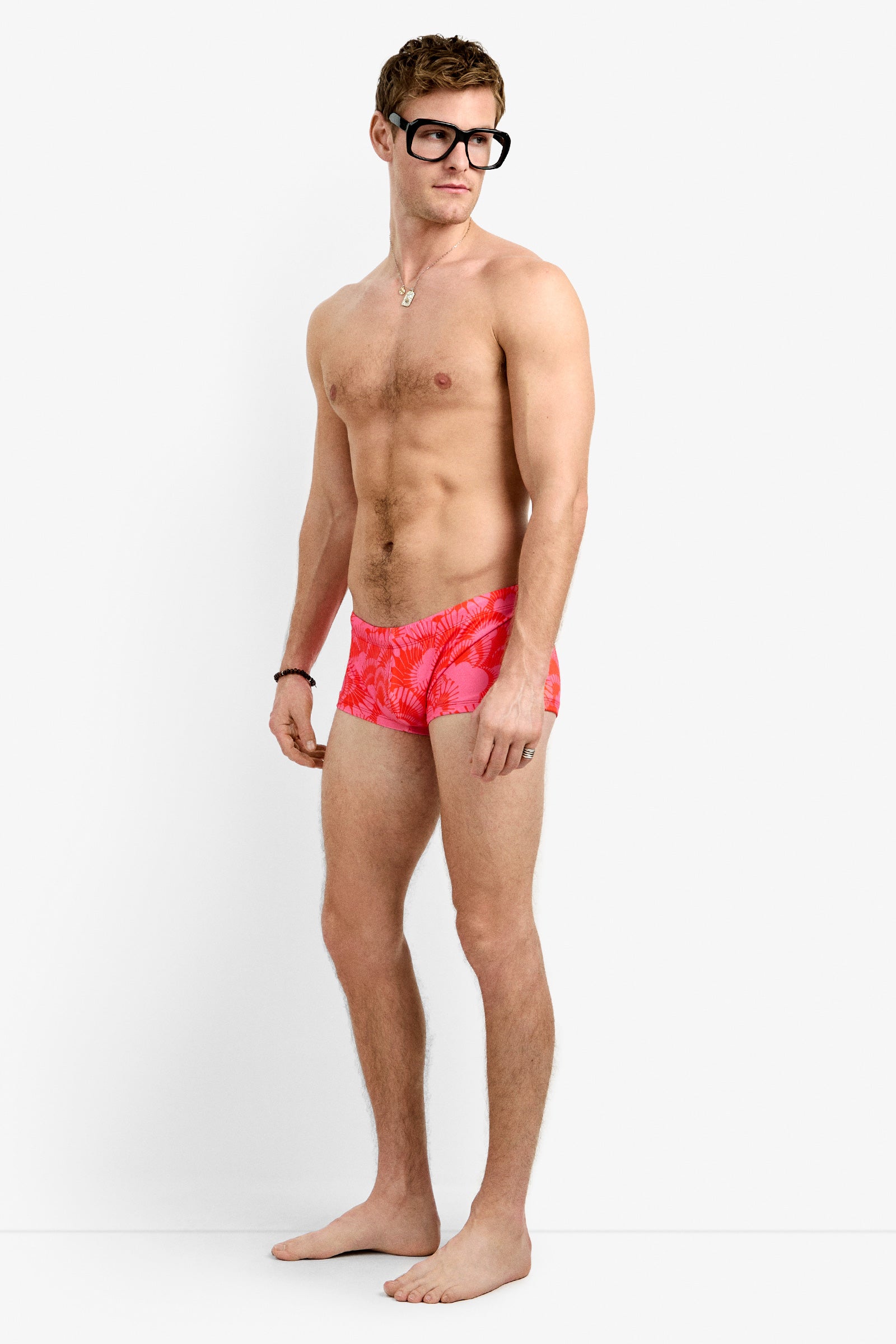 A barefoot man in large black glasses and Sorrento Swim Trunk stands against a plain white background, gazing slightly to his left.