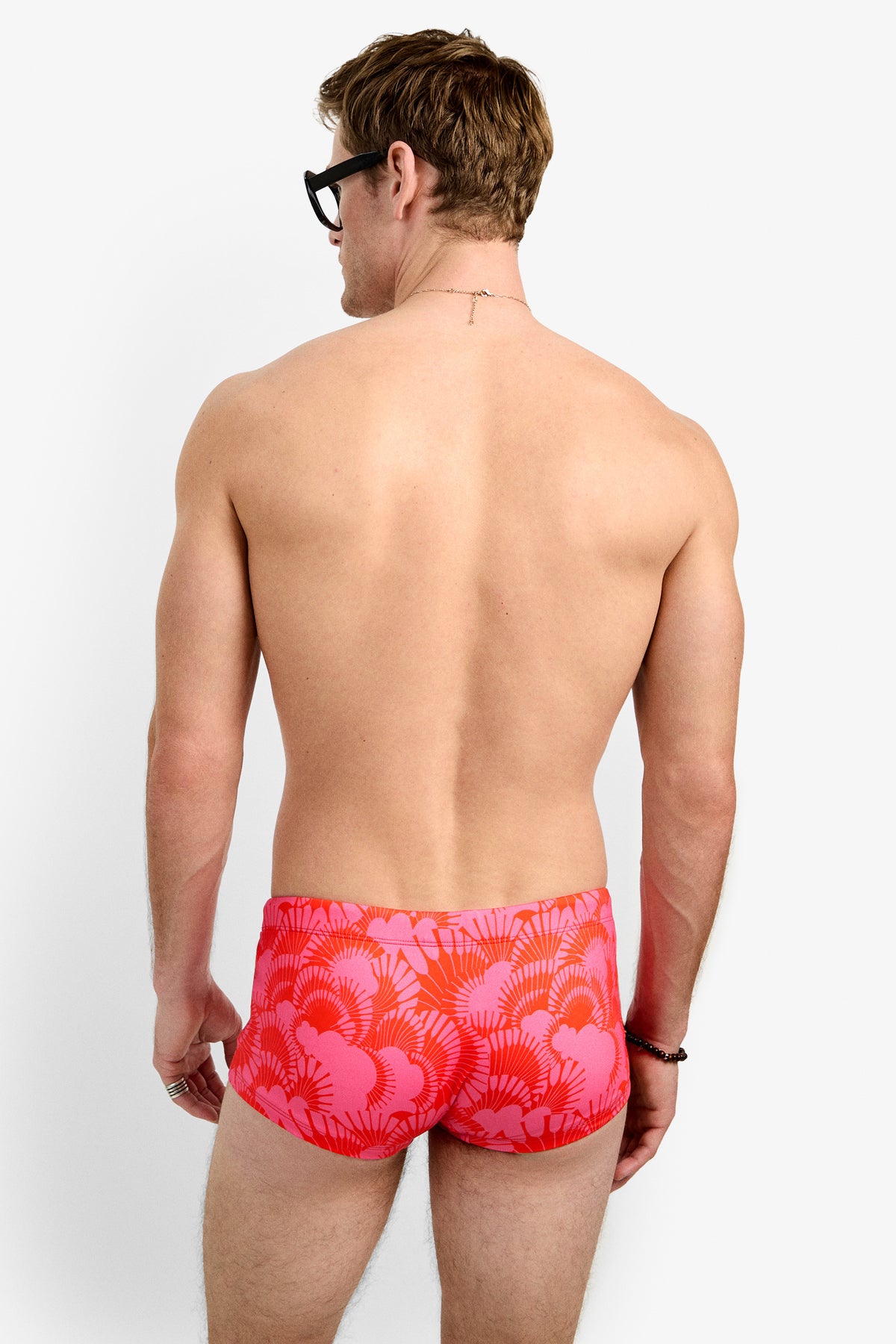 A man with short brown hair, black glasses, and red Sorrento Swim Trunk featuring a pink tropical leaf pattern stands facing away against a plain white background.