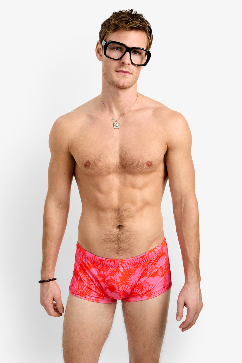 A man with large black glasses and a relaxed expression models the Sorrento Swim Trunk, featuring a bright pink pattern, along with a necklace and bracelet, against a plain white background.