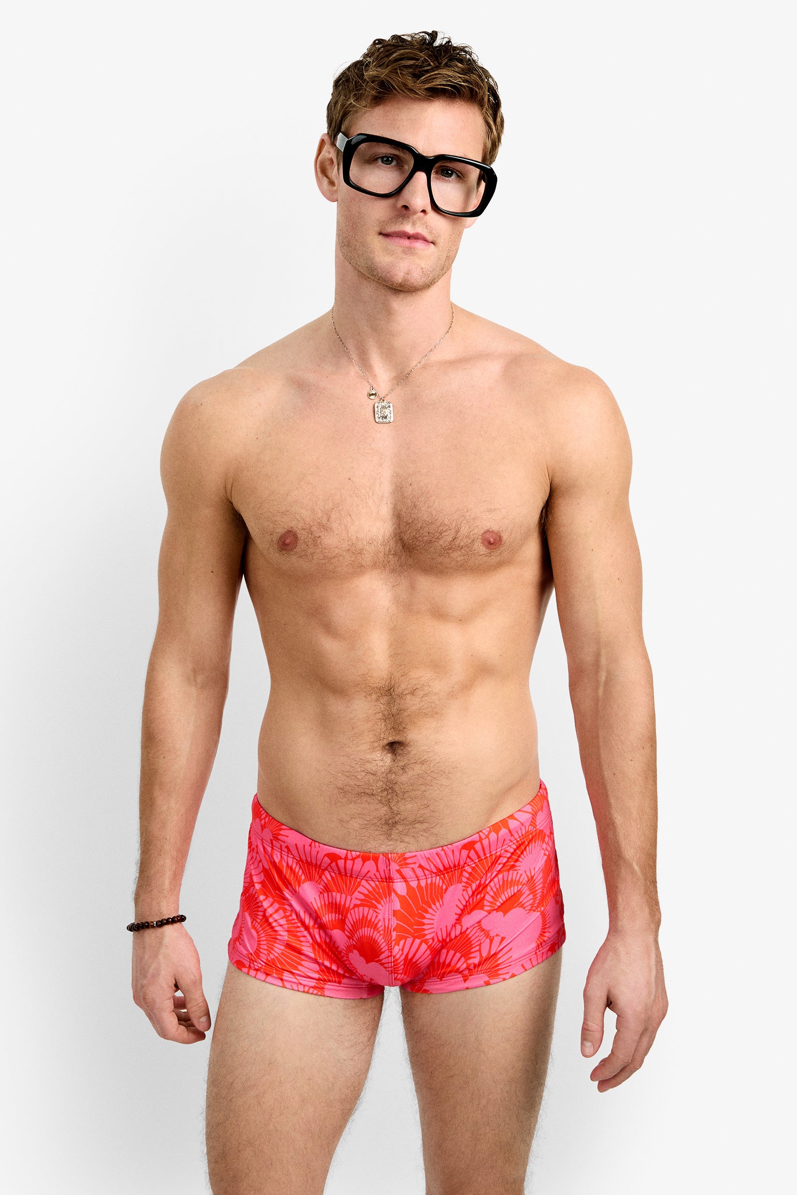 A man with large black glasses and a relaxed expression models the Sorrento Swim Trunk, featuring a bright pink pattern, along with a necklace and bracelet, against a plain white background.