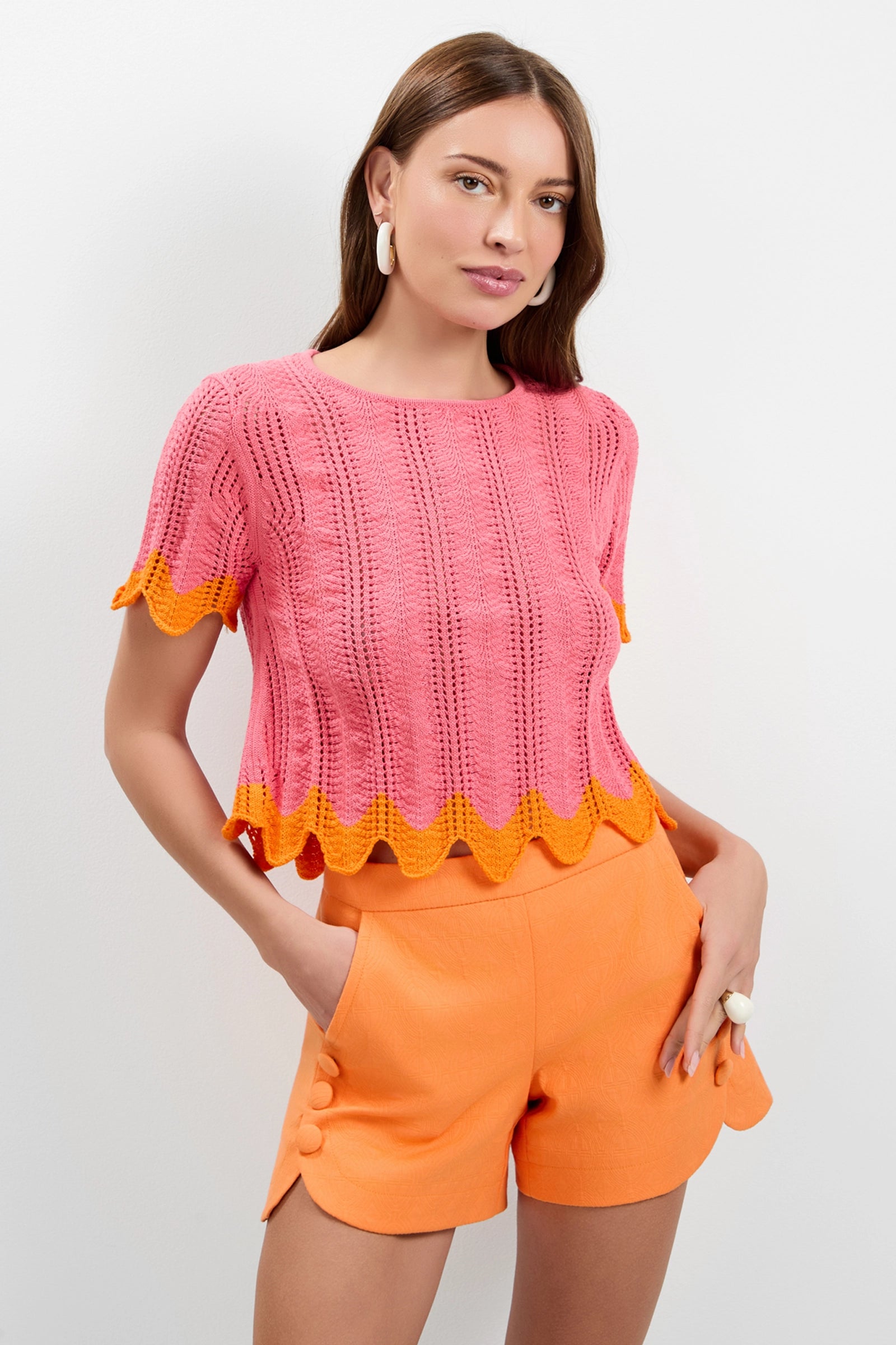 A woman models the Playa Largo Wavy Knit Top in pink with yellow scalloped edges, pairing it with orange buttoned shorts. She poses against a white background, accessorized with hoop earrings and a ring.