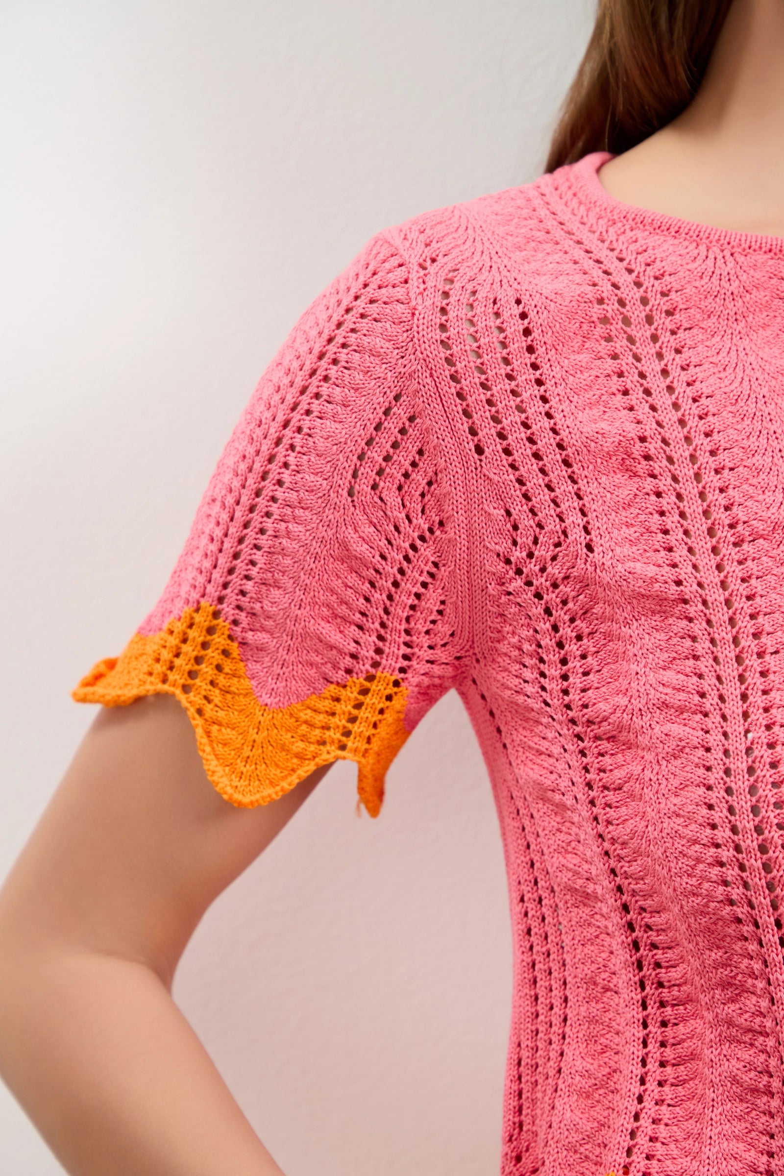 A close-up of a person wearing the Playa Largo Wavy Knit Top, featuring pink knit fabric with intricate textured patterns and short sleeves accented by an orange wavy trim.