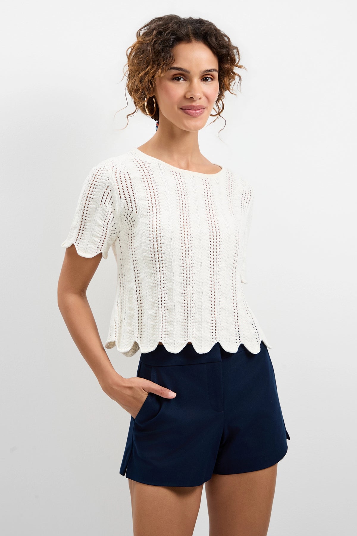 A woman with curly hair wears the Playa Largo Wavy Knit Top in white and navy blue shorts, standing against a plain white background and smiling slightly with her hands in her pockets.