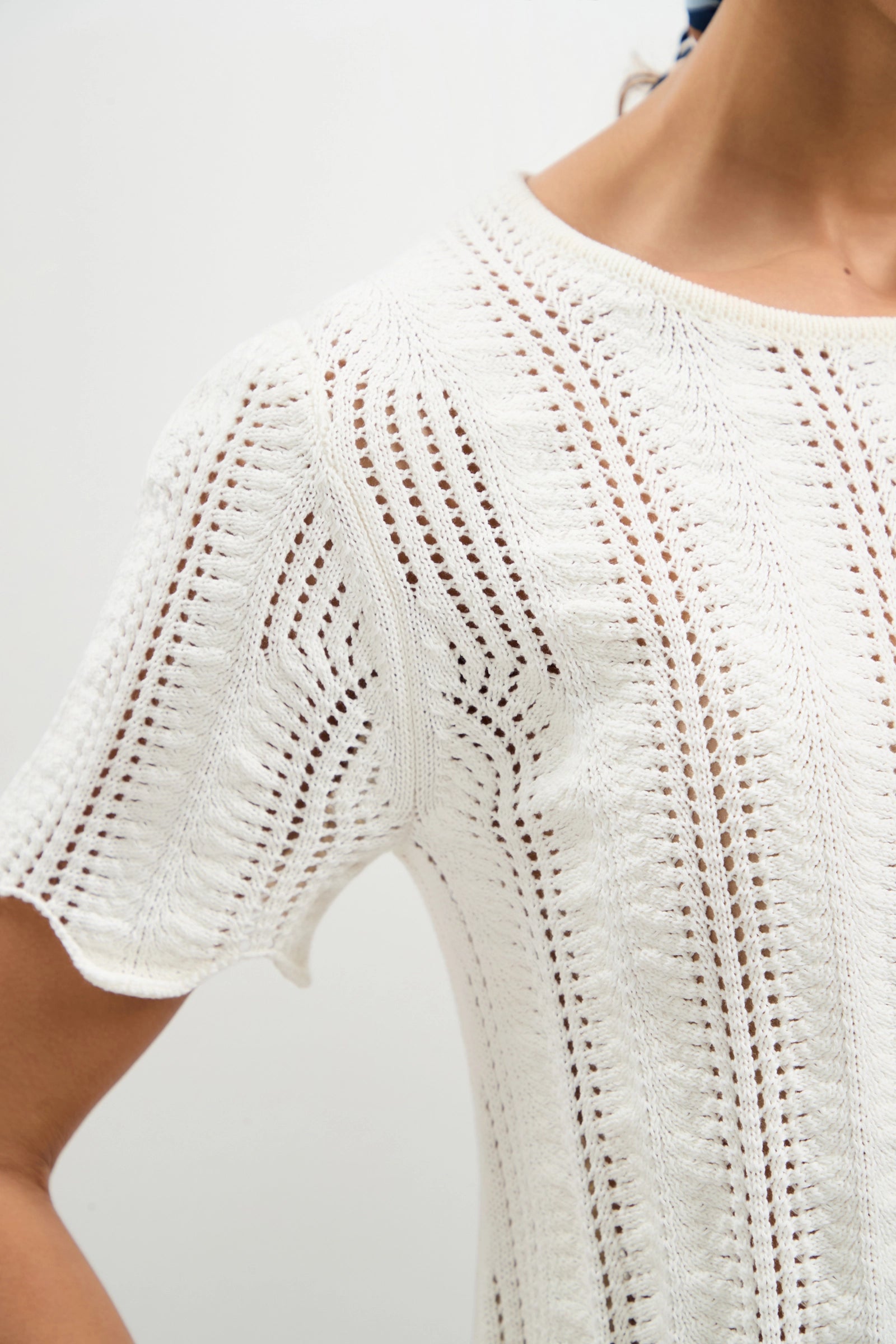 Close-up of a person wearing the Playa Largo Wavy Knit Top, a white short-sleeved design featuring intricate openwork patterns. The knit’s texture and details stand out clearly against a plain background.