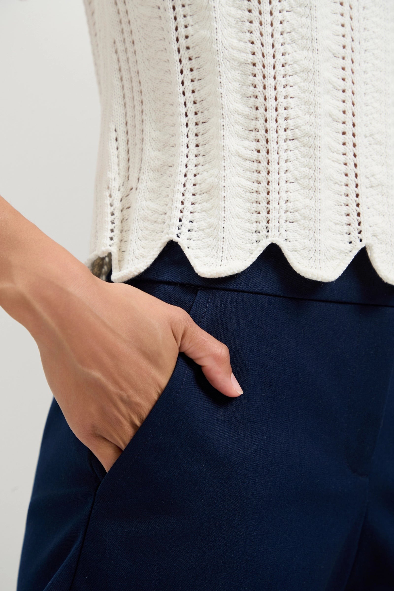A person wears the Playa Largo Wavy Knit Top, a white textured, scallop-edged top, paired with dark blue pants and has one hand casually placed in their pocket.