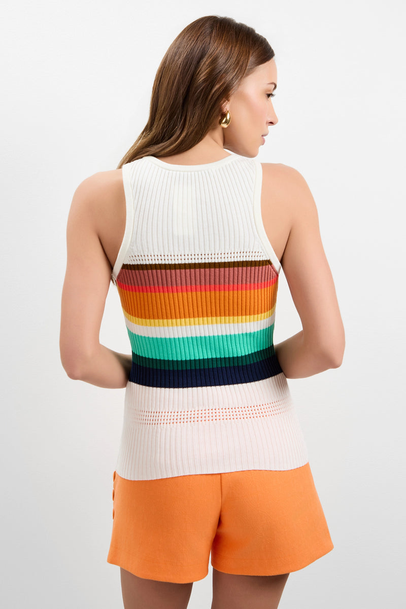 A woman with long brown hair stands against a plain light background, facing away from the camera, wearing the Violetta Tank—a sleeveless white knit top with colorful horizontal stripes on the back—and orange shorts.