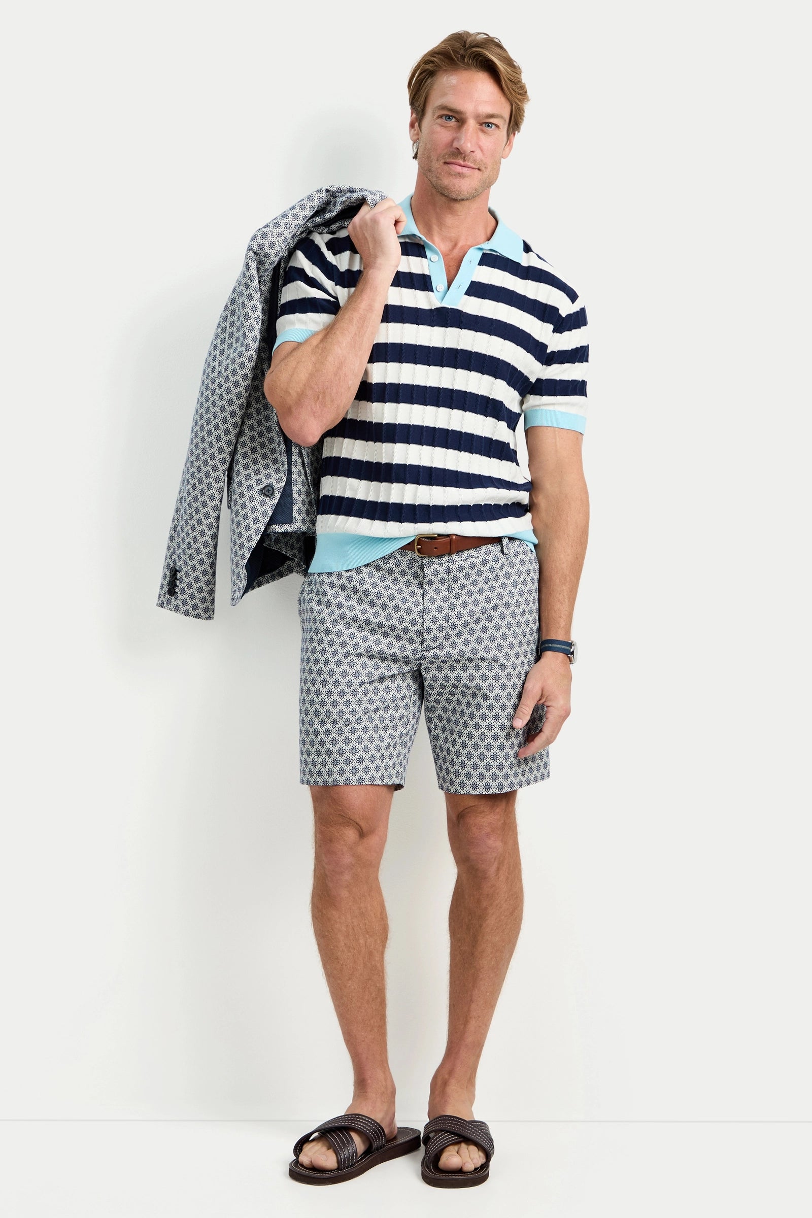 A man stands against a white background wearing the Enzo Short-Sleeve Polo, patterned shorts, a brown belt, black sandals, and holding a patterned jacket over his shoulder.