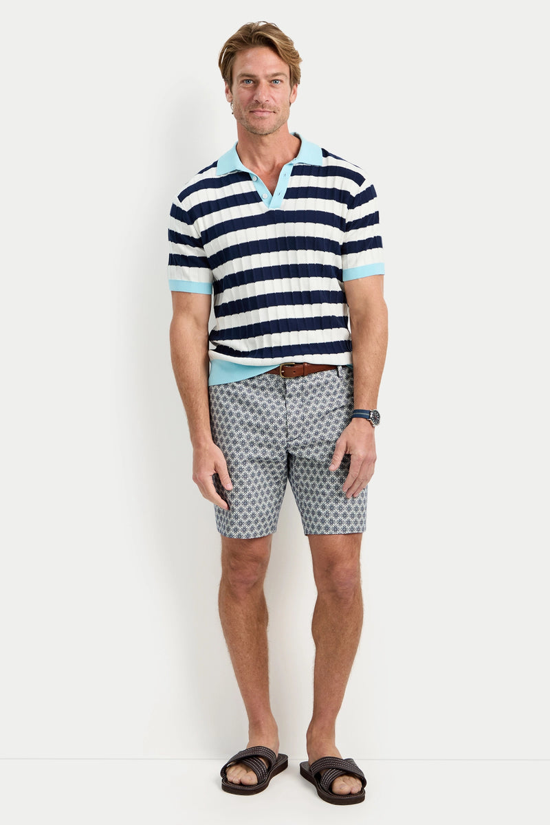 A man with short light brown hair smiles in an Enzo Short-Sleeve Polo, patterned light blue shorts, brown belt and sandals, and a black watch. He stands against a plain white background.