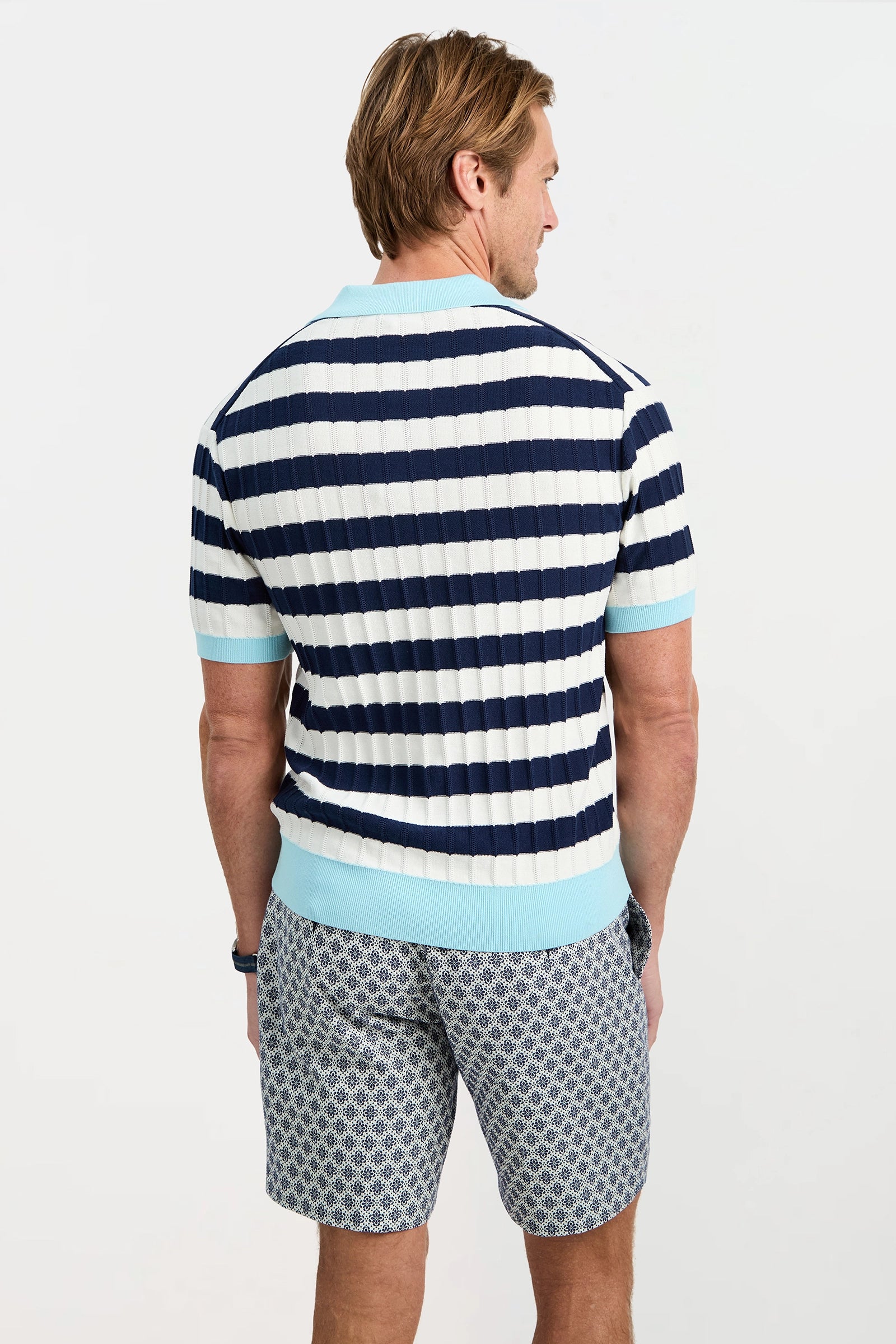 A man stands facing away, wearing the Enzo Short-Sleeve Polo in blue and white stripes with patterned shorts; his hands rest by his sides against a plain light background.