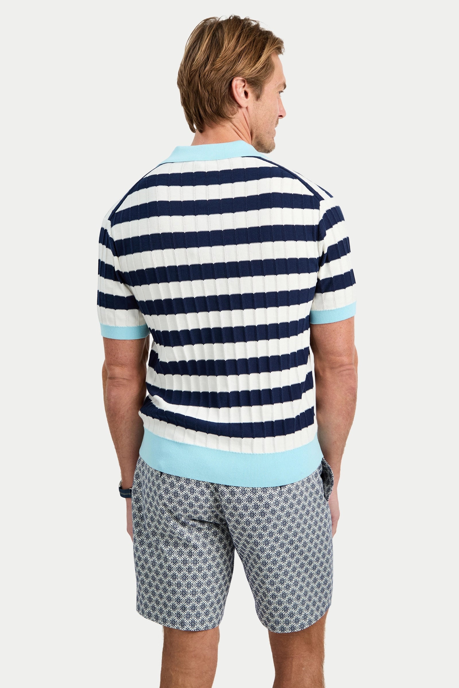 A man with light brown hair, seen from behind, wears the Enzo Short-Sleeve Polo in navy and white stripes with light blue trim, paired with patterned blue and white shorts.