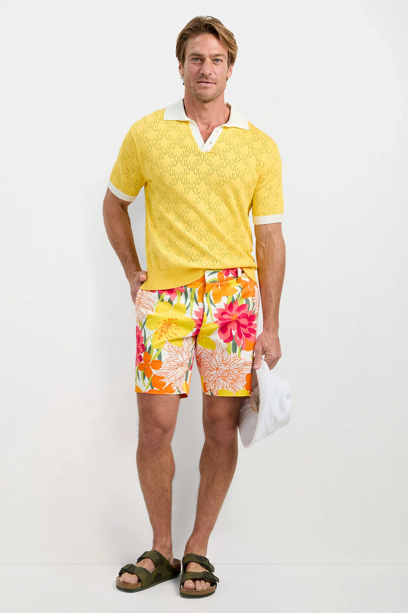 A man stands against a white background wearing Lawrence Short - Como Herringbone, a yellow patterned polo shirt, dark sandals, and holding a white cap by his side.