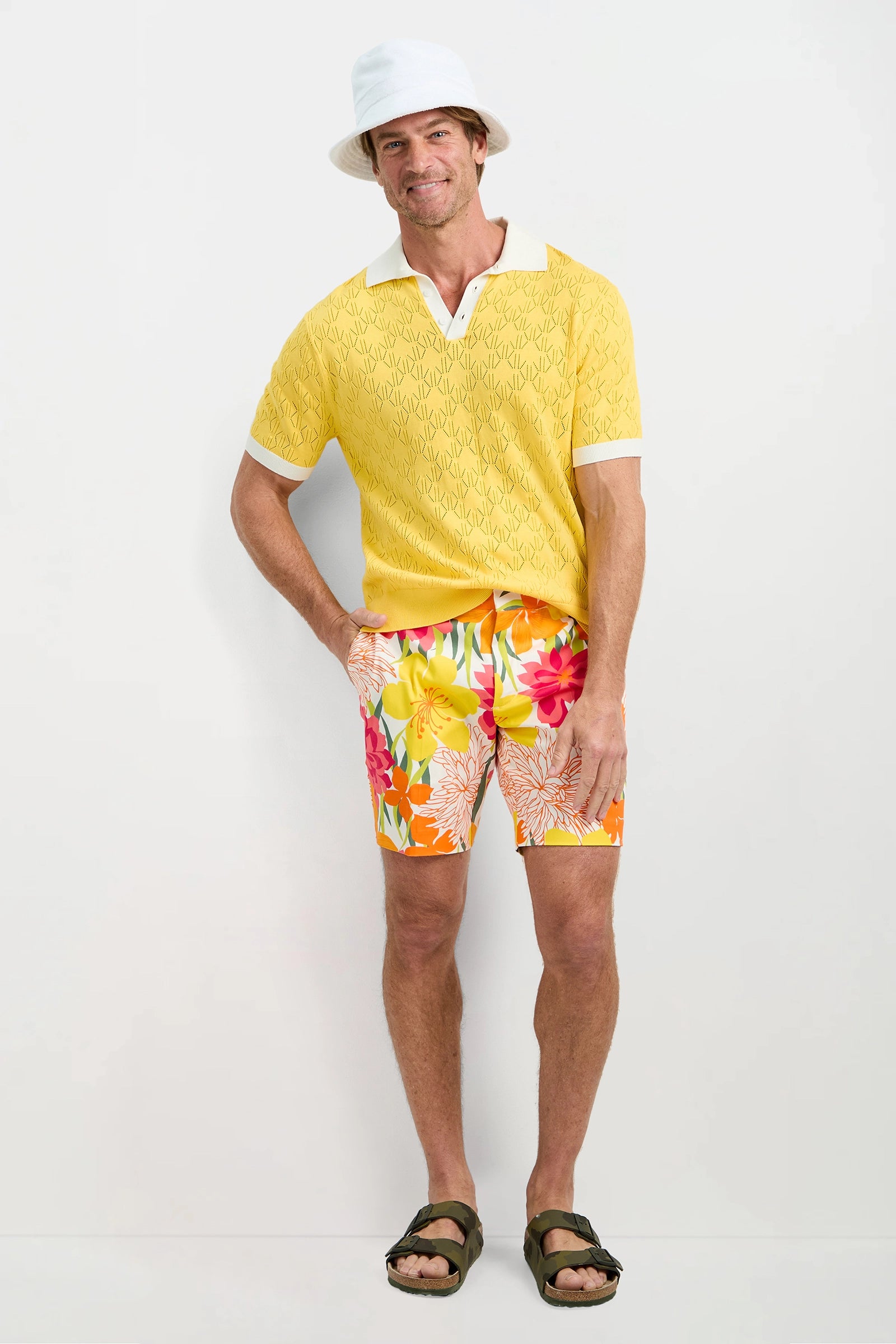 A man stands against a white wall wearing the Luca Short-Sleeve Polo in yellow, a white bucket hat, colorful floral shorts, and dark green sandals. He smiles with one hand in his pocket and the other resting by his side.