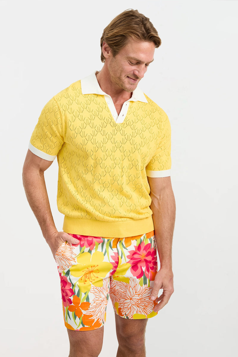 A man wears the Luca Short-Sleeve Polo in yellow with a white collar and cuffs, paired with colorful floral shorts. He smiles while looking down against a plain light background.