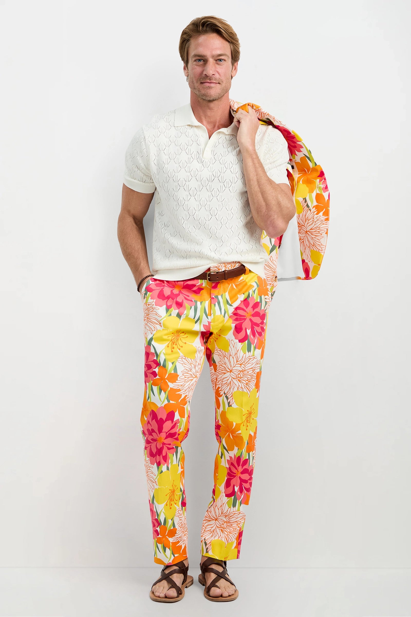 A man stands against a white background wearing the Luca Short-Sleeve Polo, vibrant floral pants in orange, yellow, and pink, brown sandals, and holding a matching floral jacket over his shoulder.