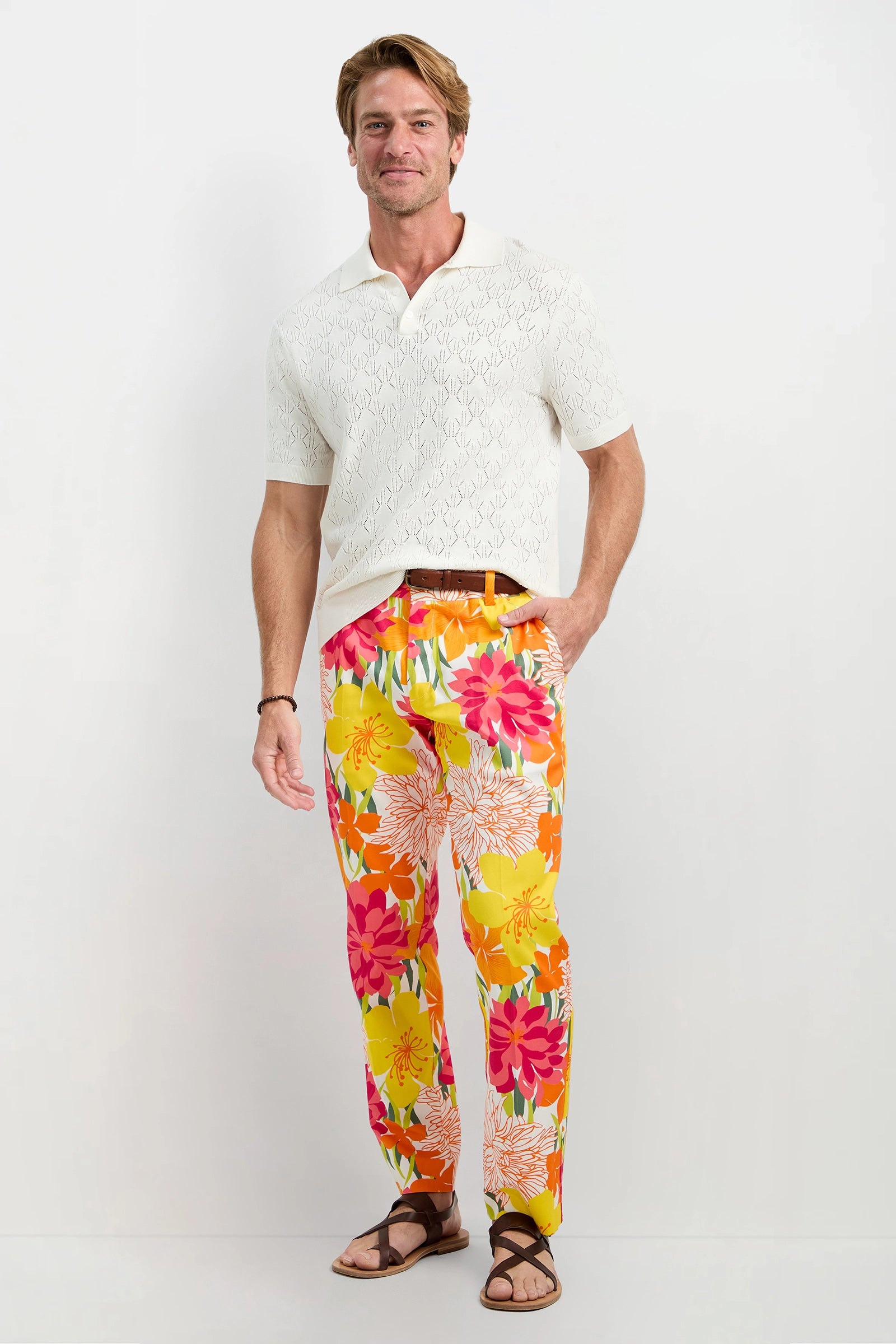 A man smiles with one hand in his pocket, modeling the white Luca Short-Sleeve Polo, bright floral pants with pink, orange, and yellow flowers, a brown belt, and brown sandals against a plain white background.