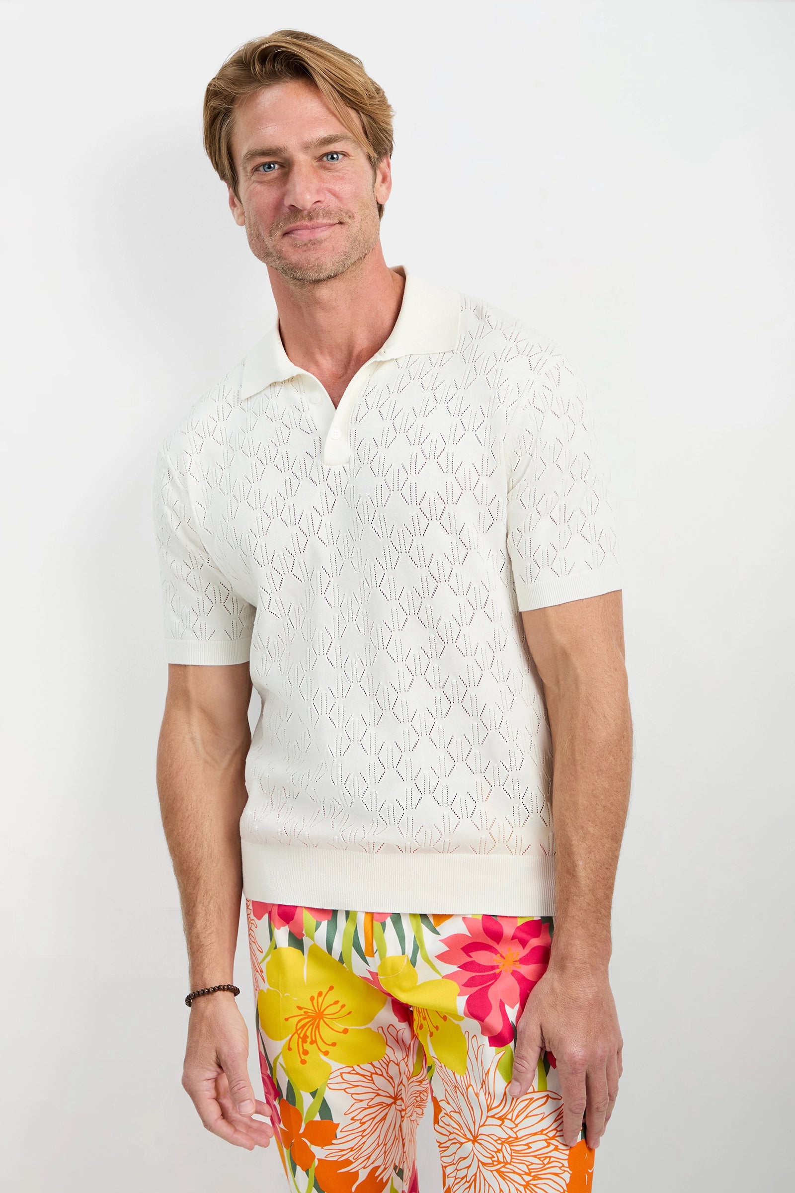 A man stands against a white background, smiling with one arm relaxed, wearing the Luca Short-Sleeve Polo in white with a subtle pattern and vibrant floral pants in orange, yellow, pink, and red tones.