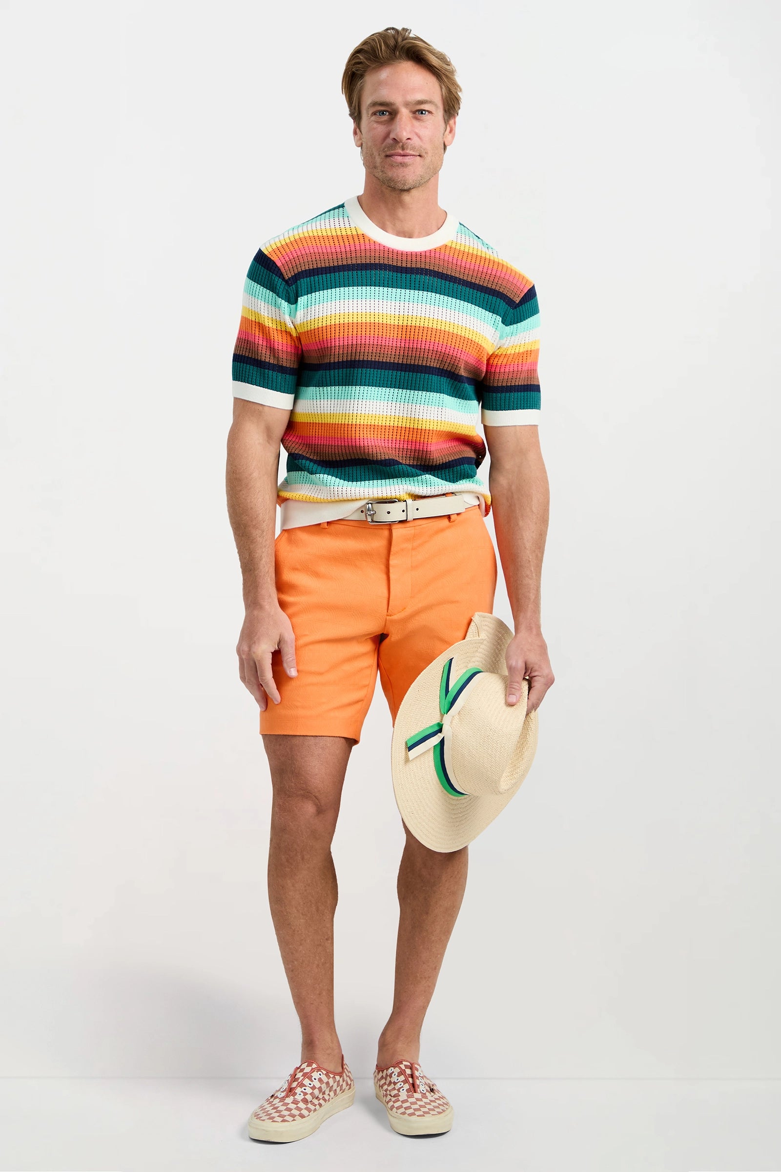 A man stands against a plain background wearing the Carlos Short-Sleeve Mesh Crewneck, orange shorts, woven shoes, and holding a straw hat with a green and orange band in his left hand.