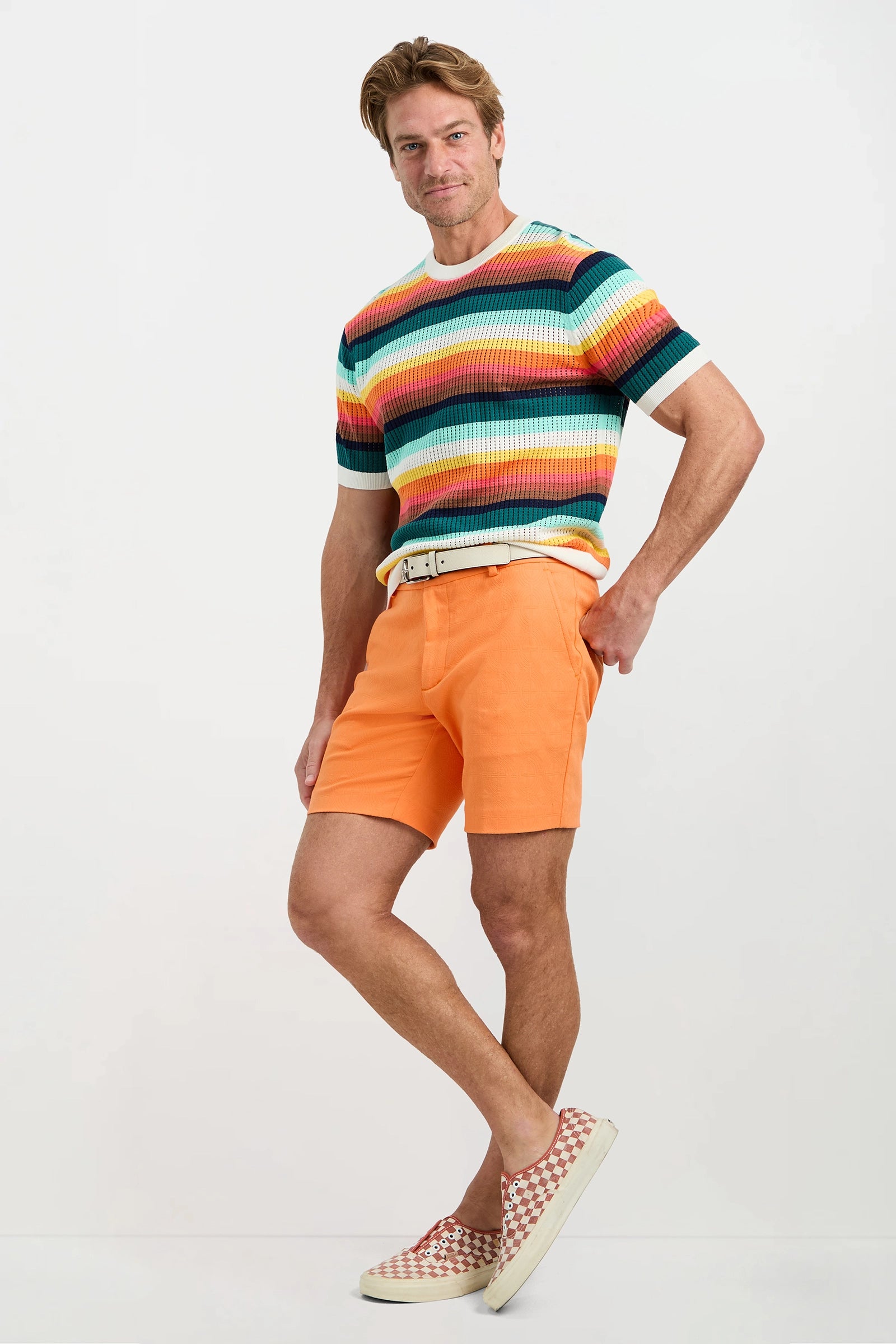 A man stands confidently against a plain background wearing the Carlos Short-Sleeve Mesh Crewneck, orange shorts with a white belt, and checkered slip-on shoes. One hand rests on his hip as he smiles slightly.