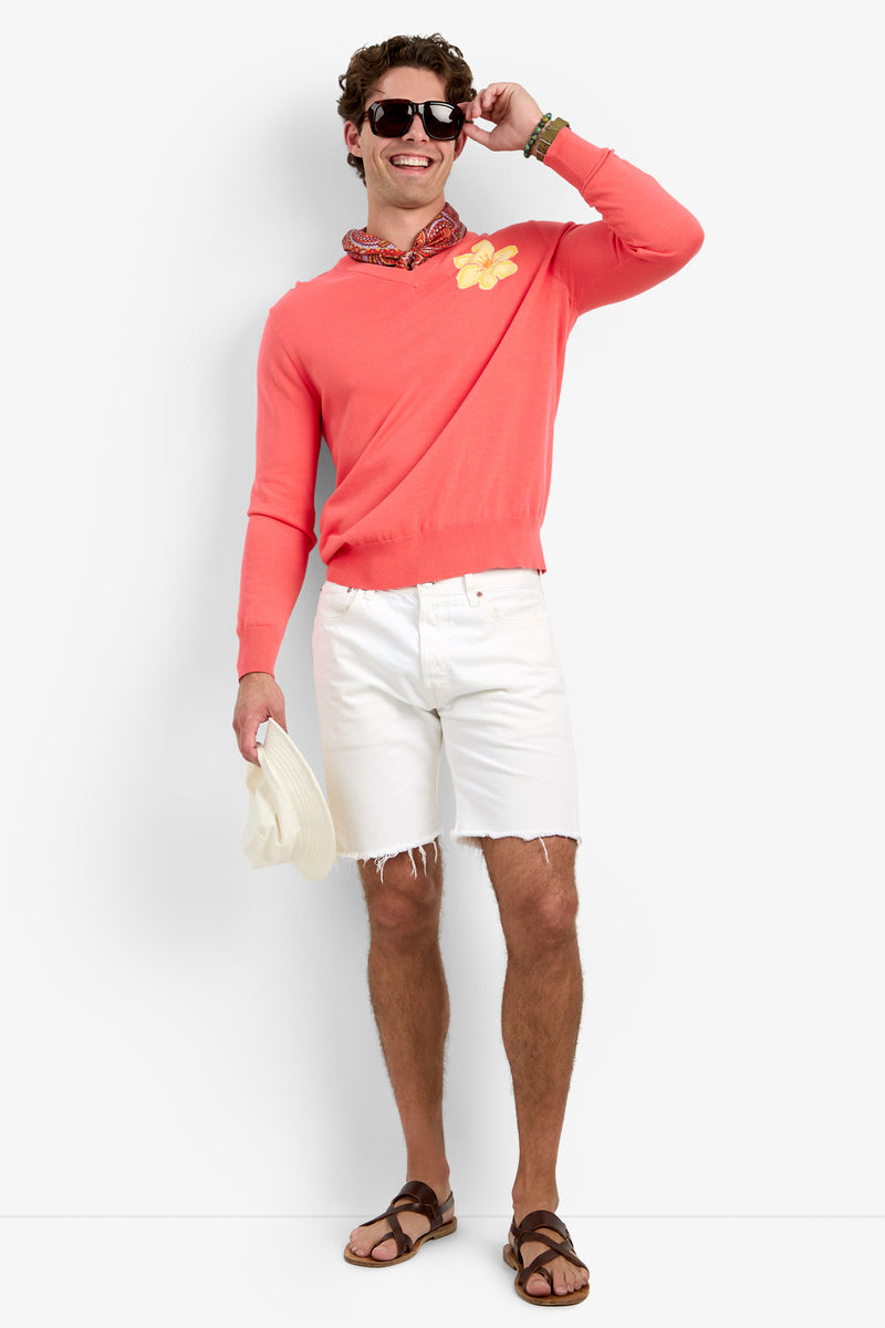 A man smiles against a plain background, wearing the Divino V-Neck Sweater in coral with a flower pin, white frayed shorts, brown sandals, sunglasses, a patterned scarf around his neck, and holding a white hat.
